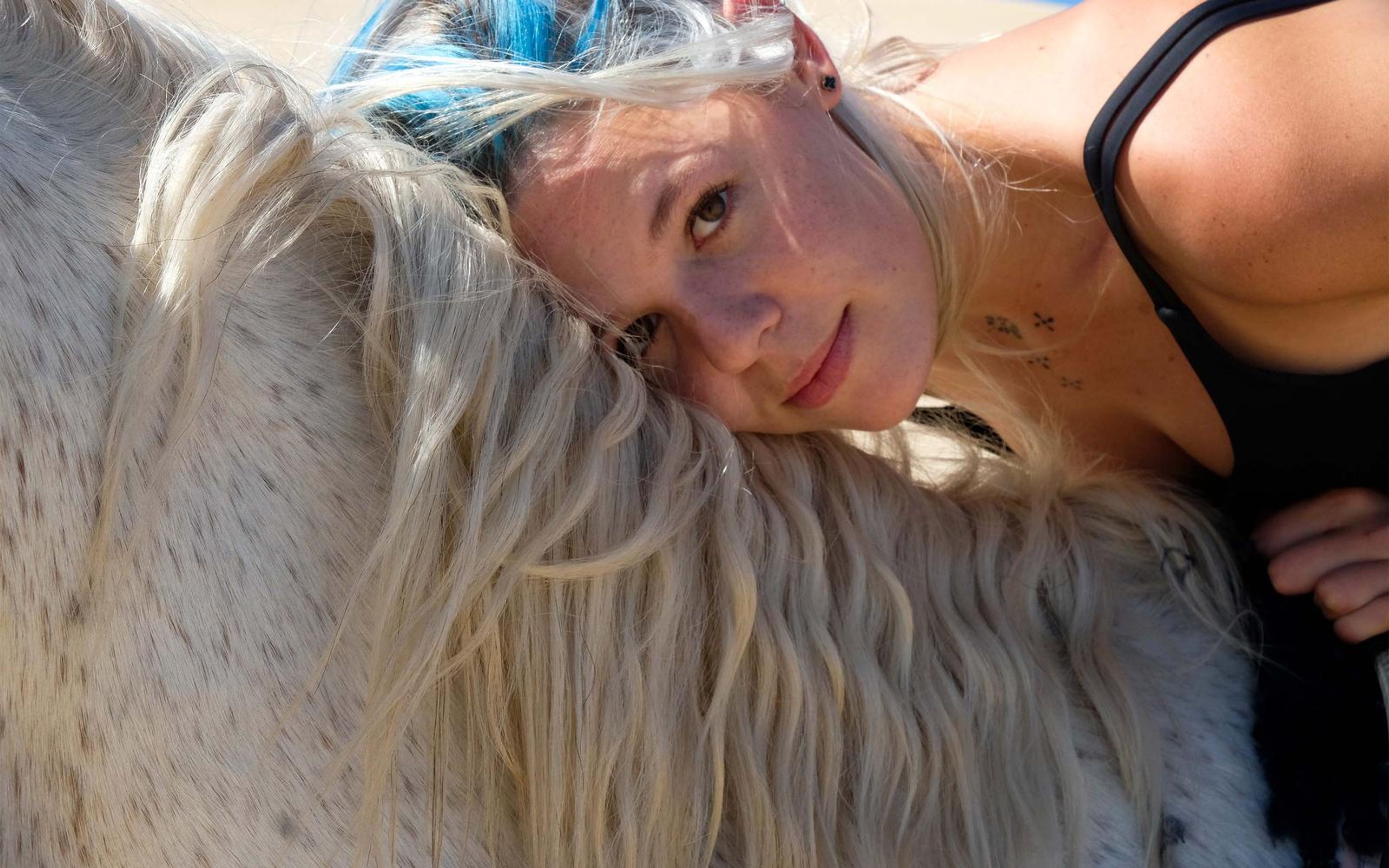 Woman with blonde and blue hair resting her cheek on a white horse's mane, smiling toward camera, beach light