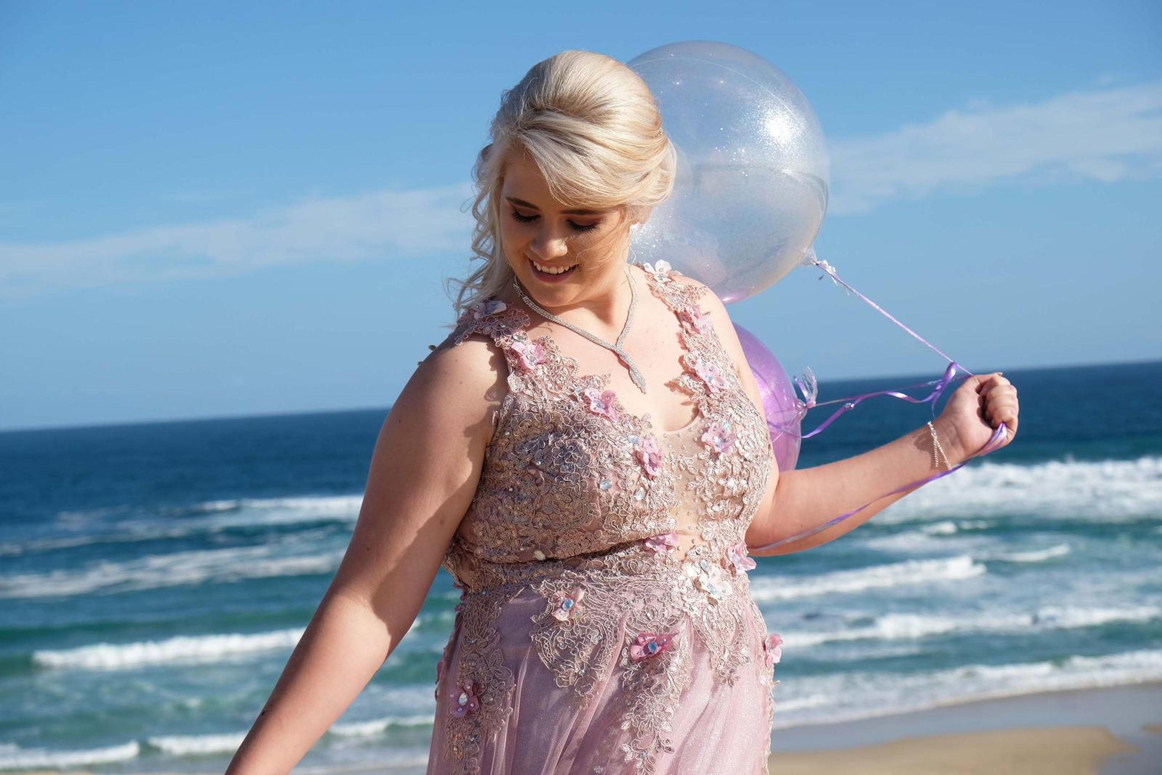 Smiling in a pink beaded gown holding balloons by the sea
