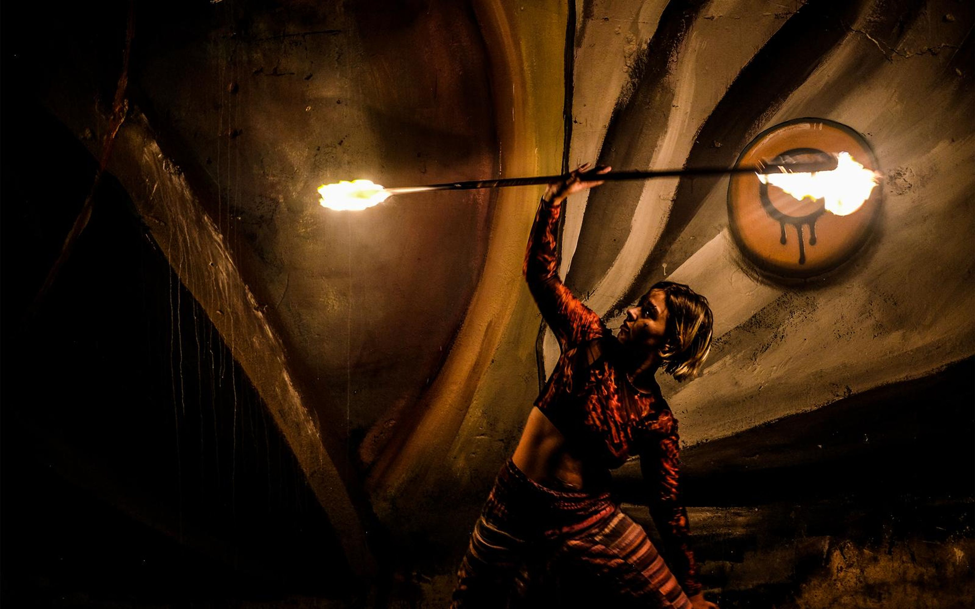 Performer with one arm raised holding a burning staff across the frame, emblem on the wall behind