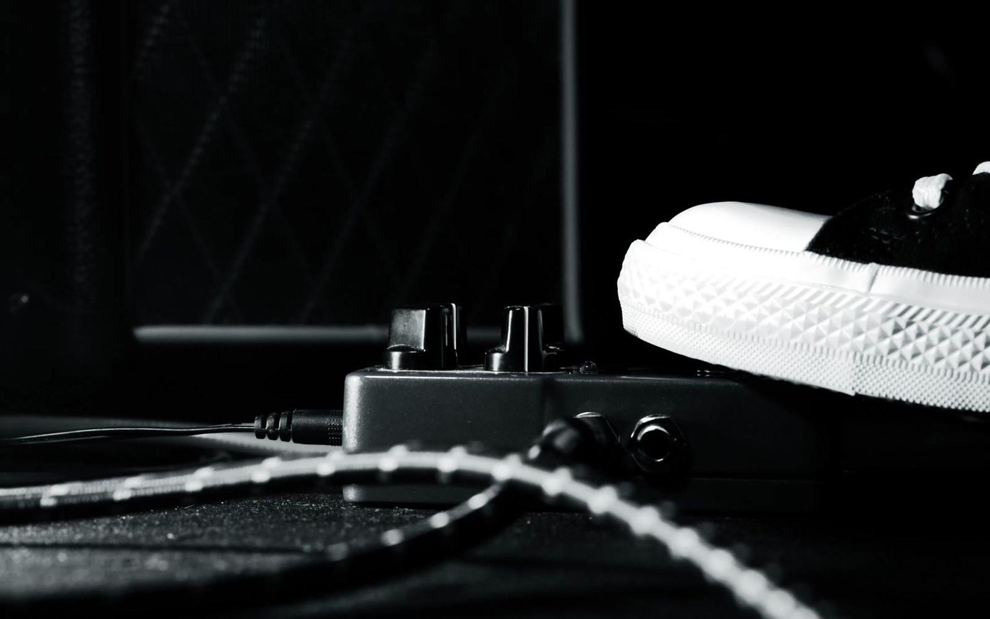 Black and white sneaker pressing an effects pedal among cables on a dark floor