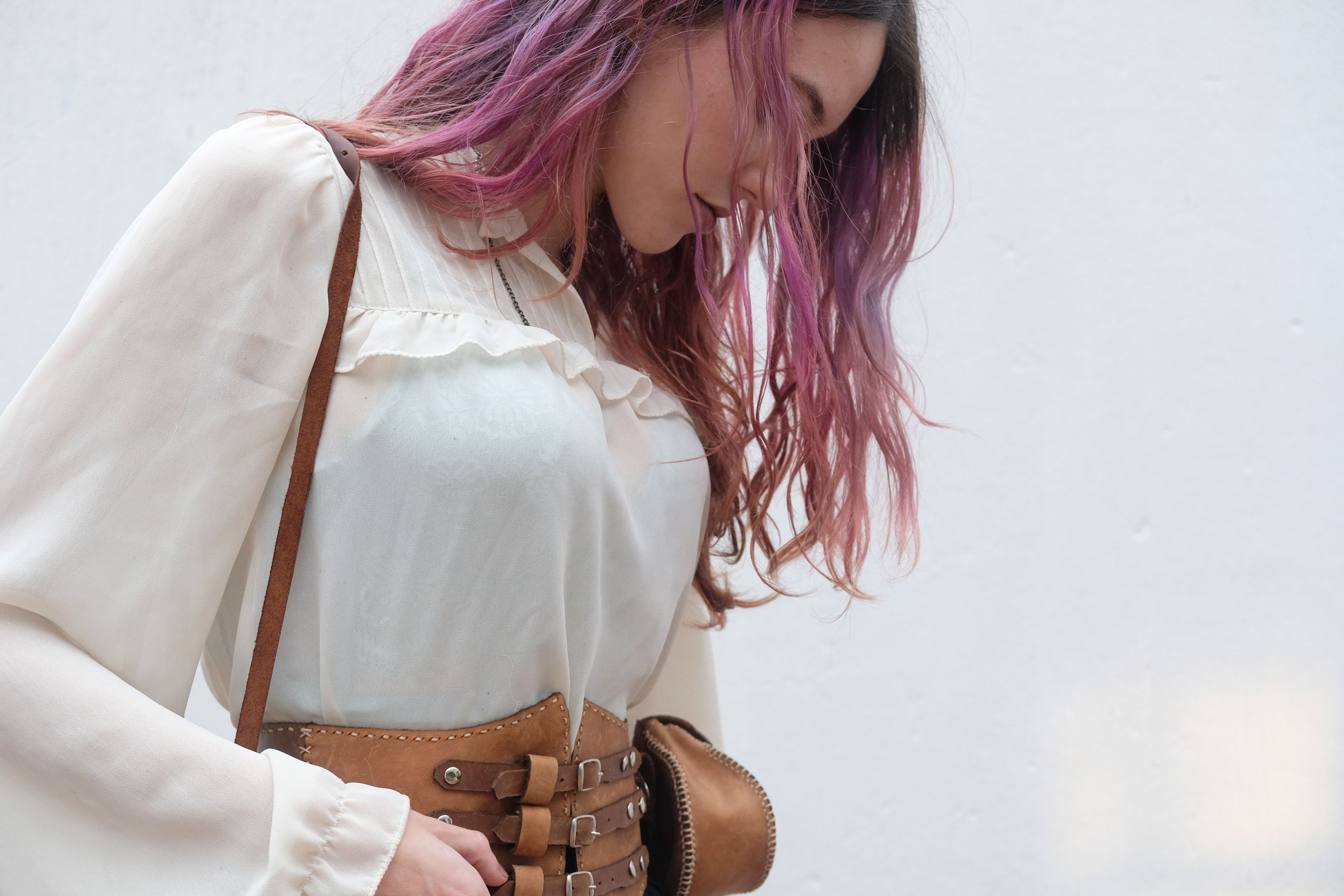 Woman in a cream blouse looking down, with a leather corset belt and a thin shoulder strap