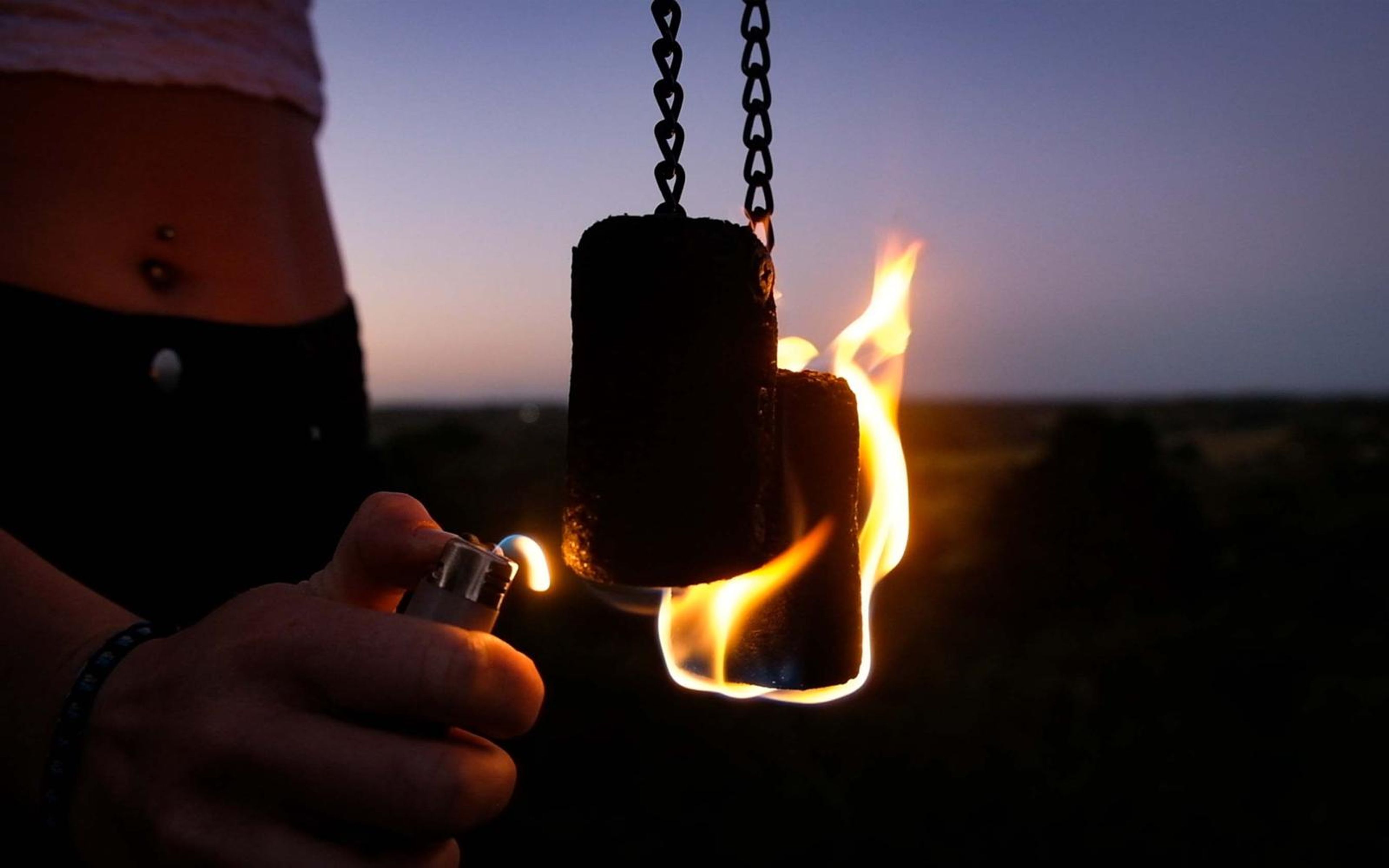Close crop of a hand holding a lighter beside a burning fire prop on chains, dark horizon behind