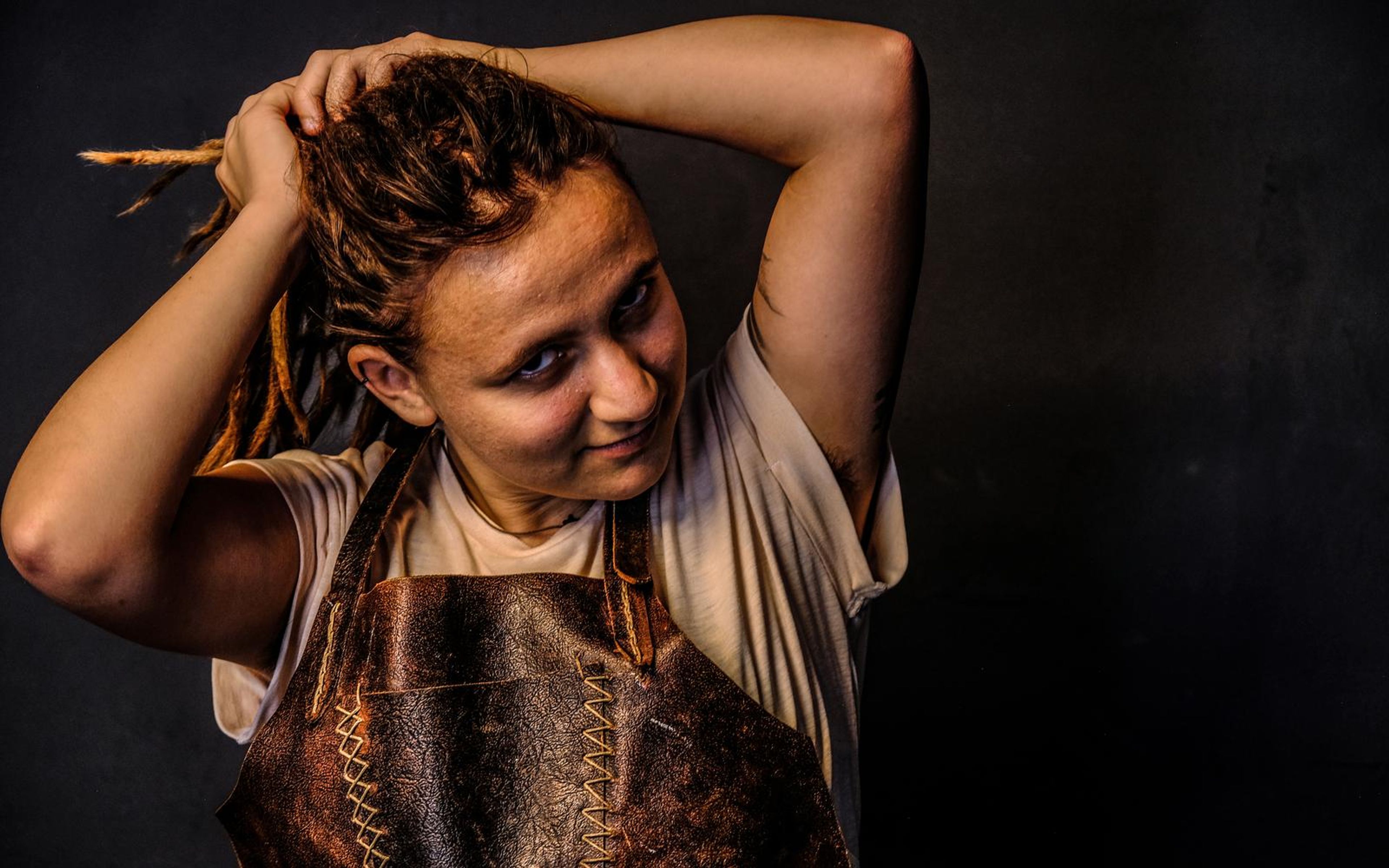 Woman in a leather apron with both hands in her dreadlocks, glancing sideways in warm light