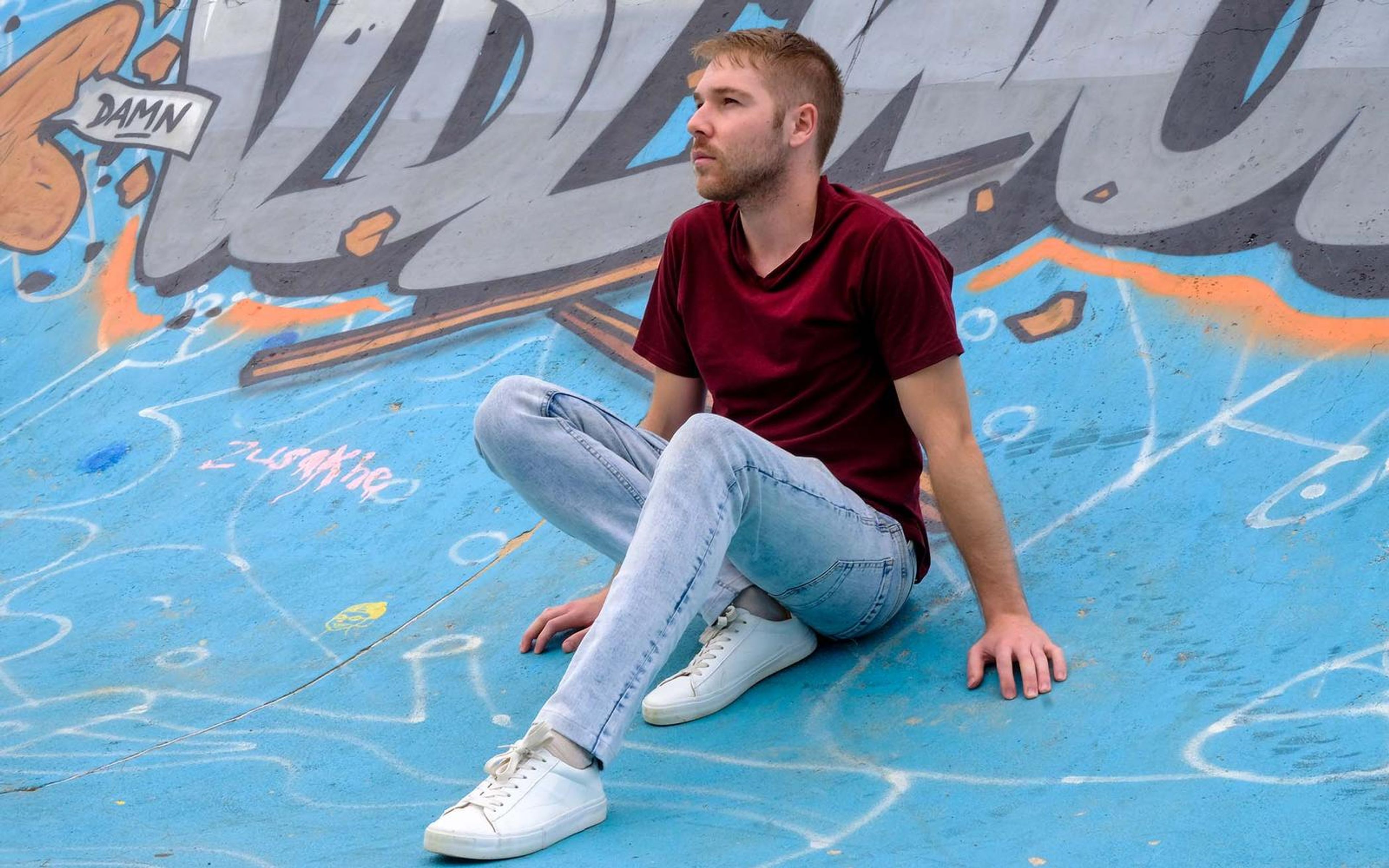 Man sitting on blue graffiti-covered ground, leaning back on one hand, looking away, white sneakers