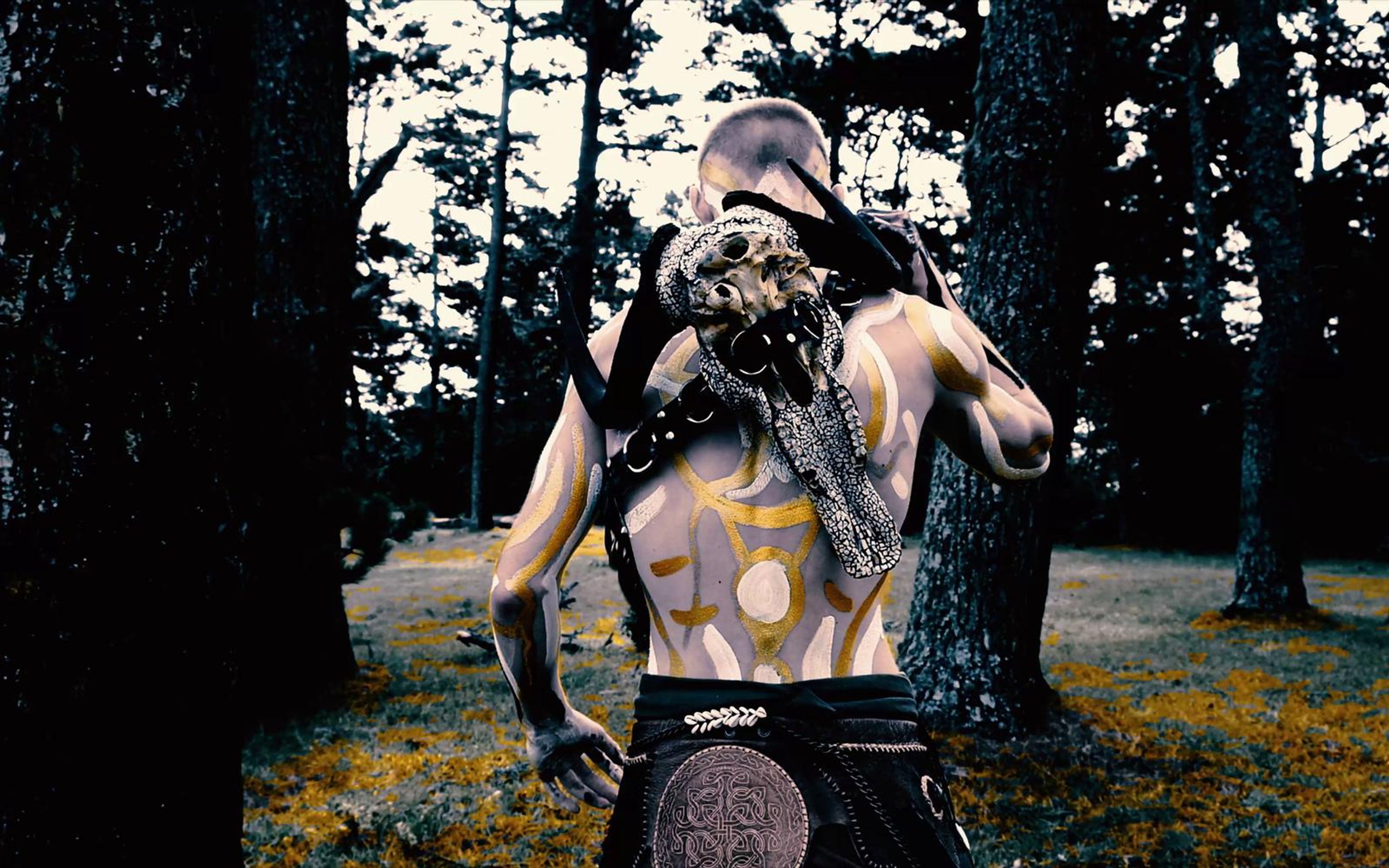 Performer standing in a clearing with a skull over his face and arms hanging, golden flowers in the foreground