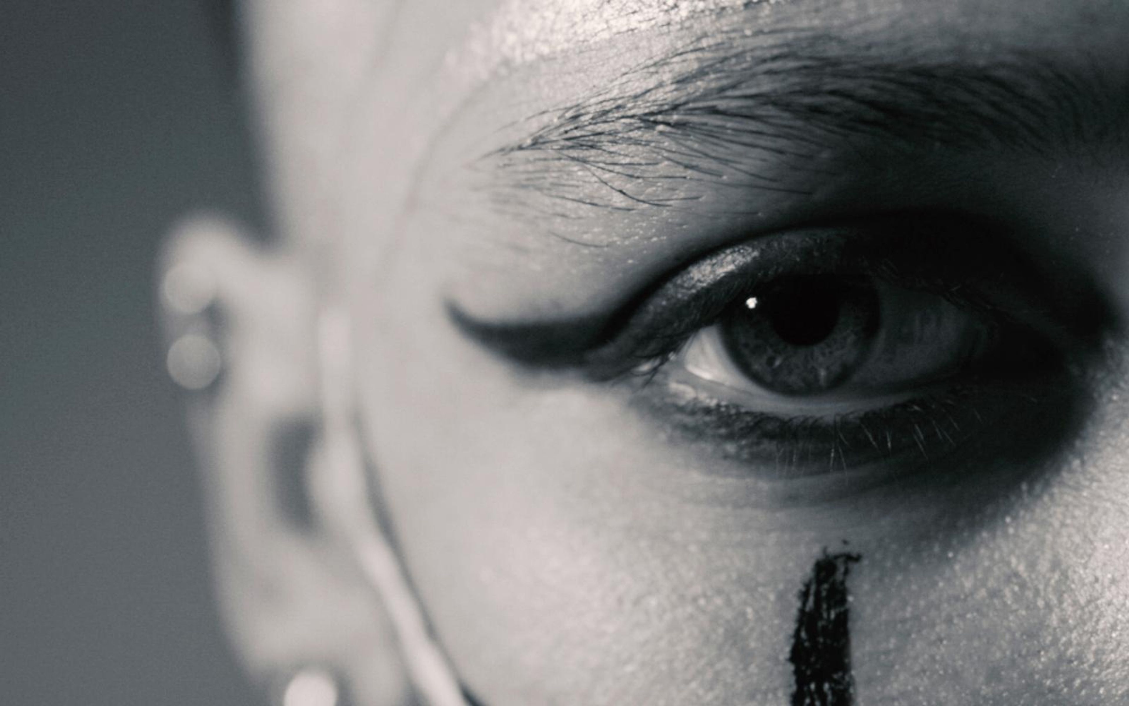 Black and white extreme close view of an eye with winged liner and a black streak painted down the cheek, shaved head