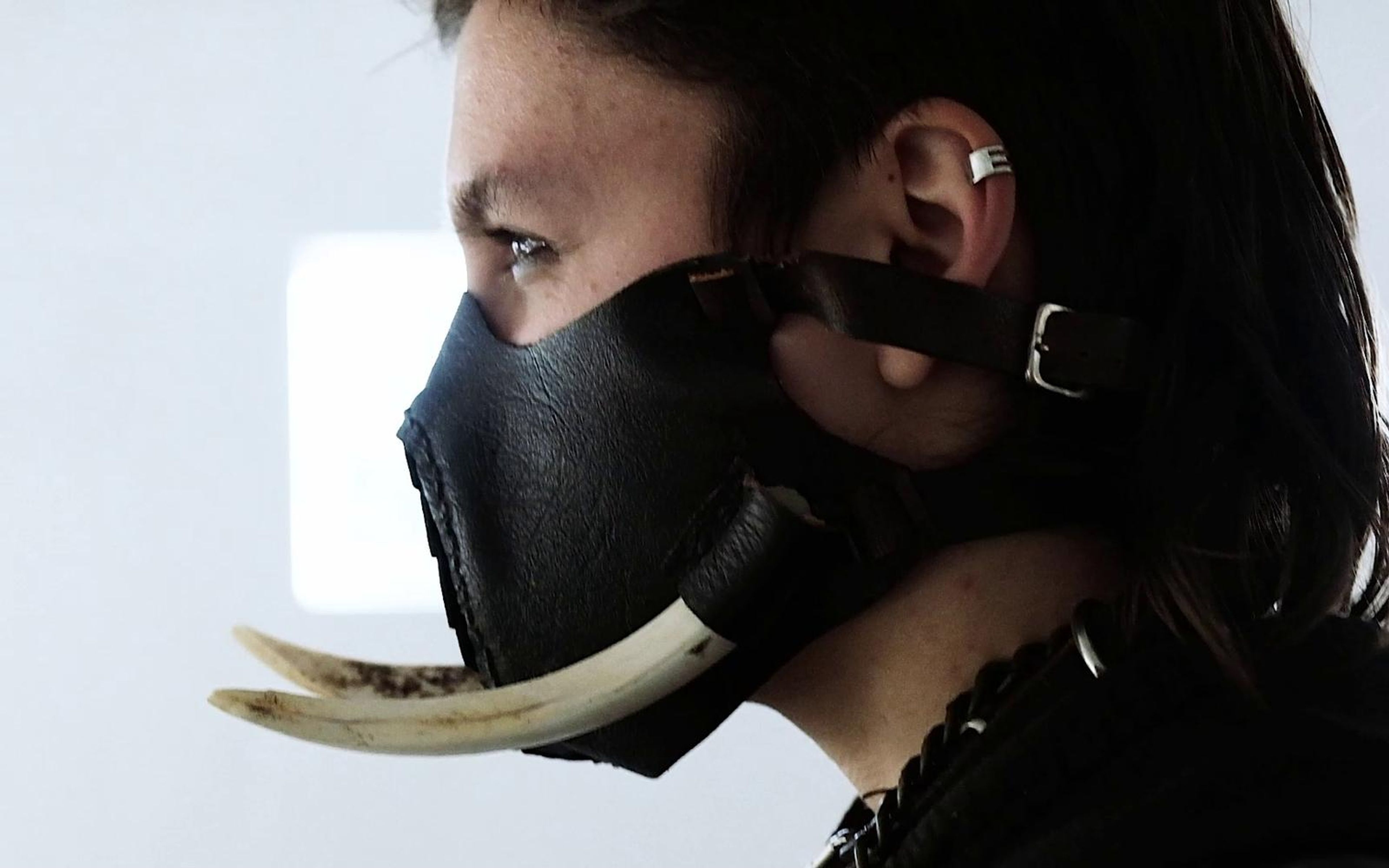 Side profile of the warrior in a black leather mask with a long curved tusk, white wall behind