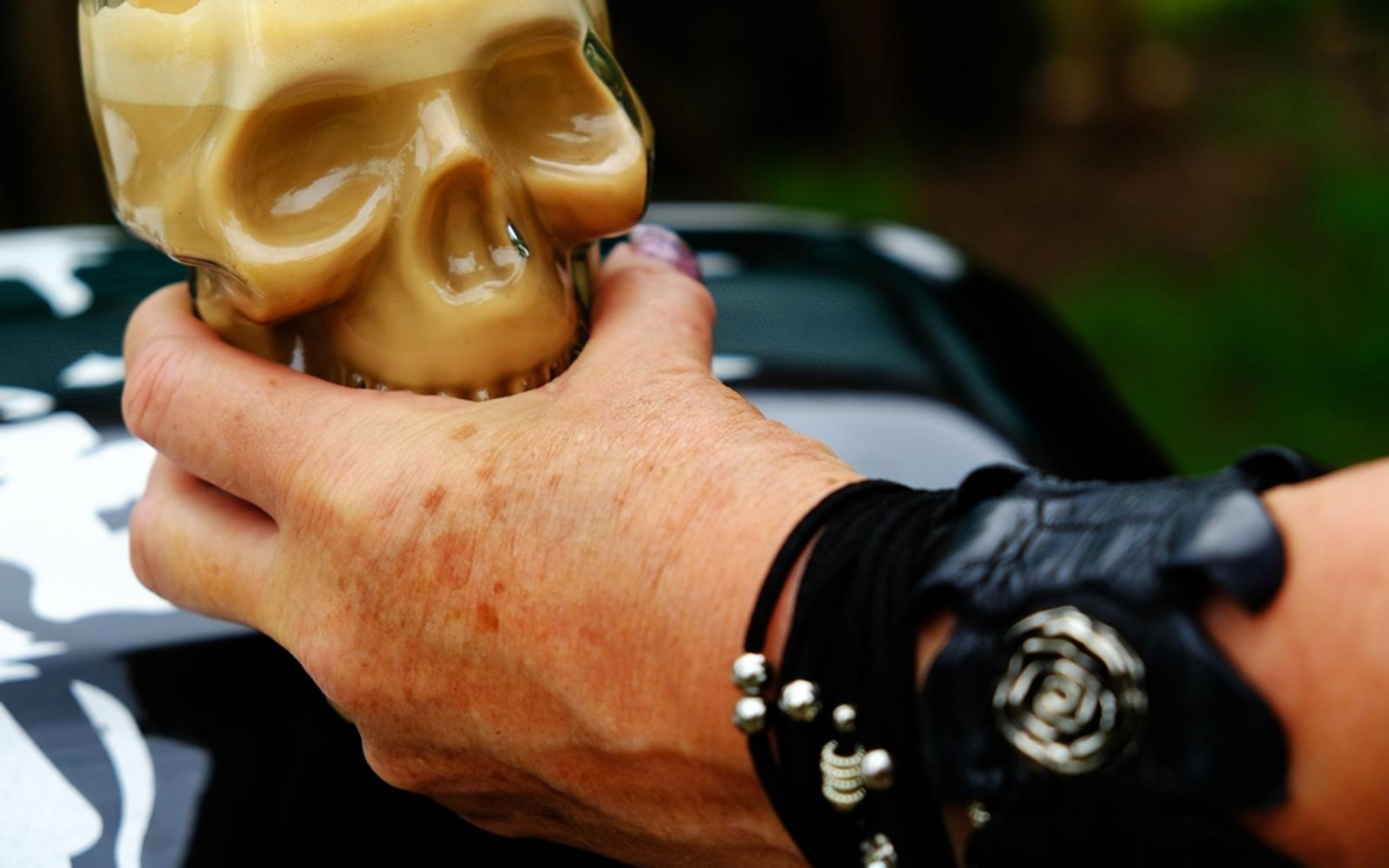 Hand holding a skull-shaped drink holder, studded black wristband and a rose detail on the cuff
