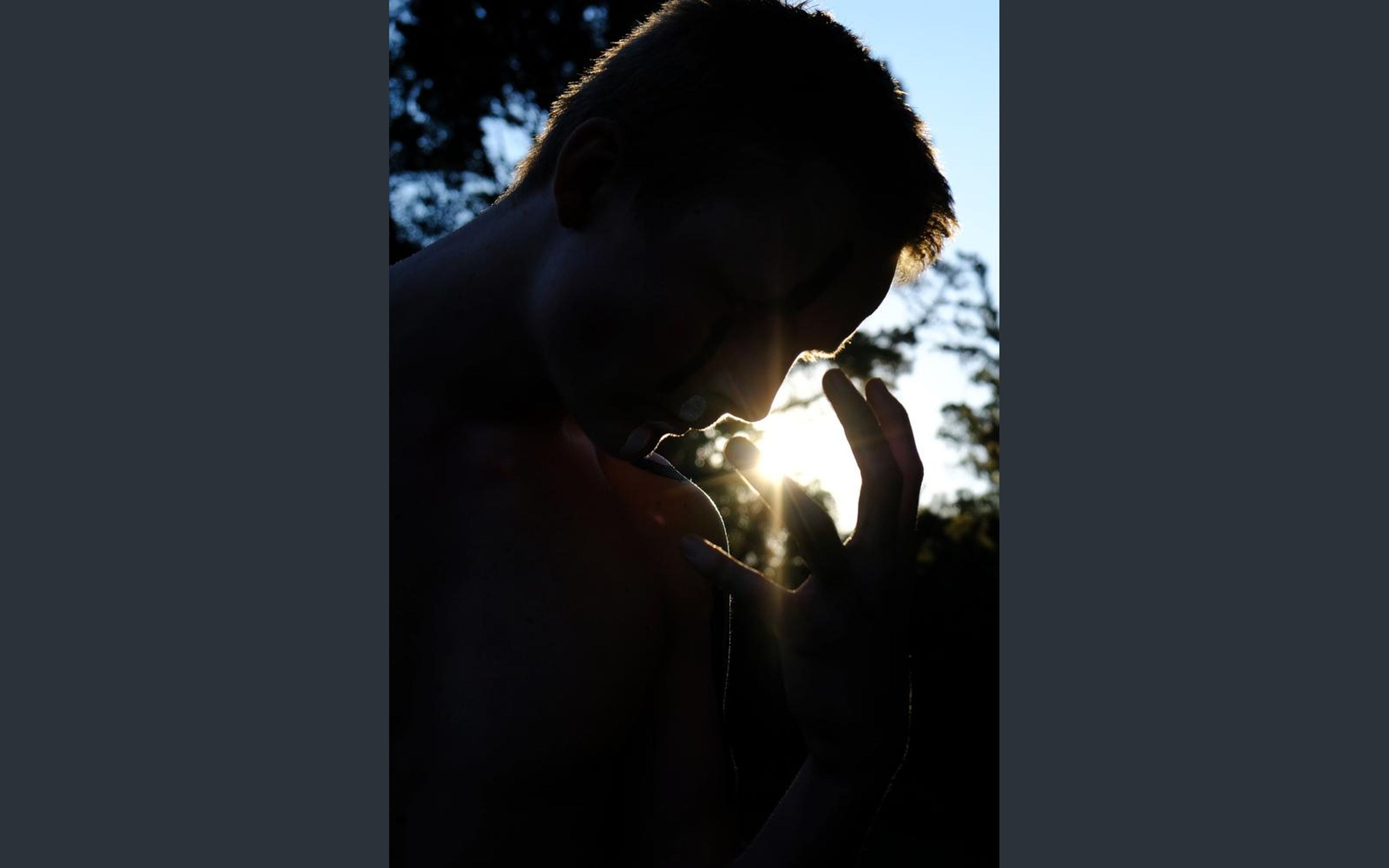 Silhouette of a performer in profile with a hand raised to the face, backlit by the low sun through pine trees