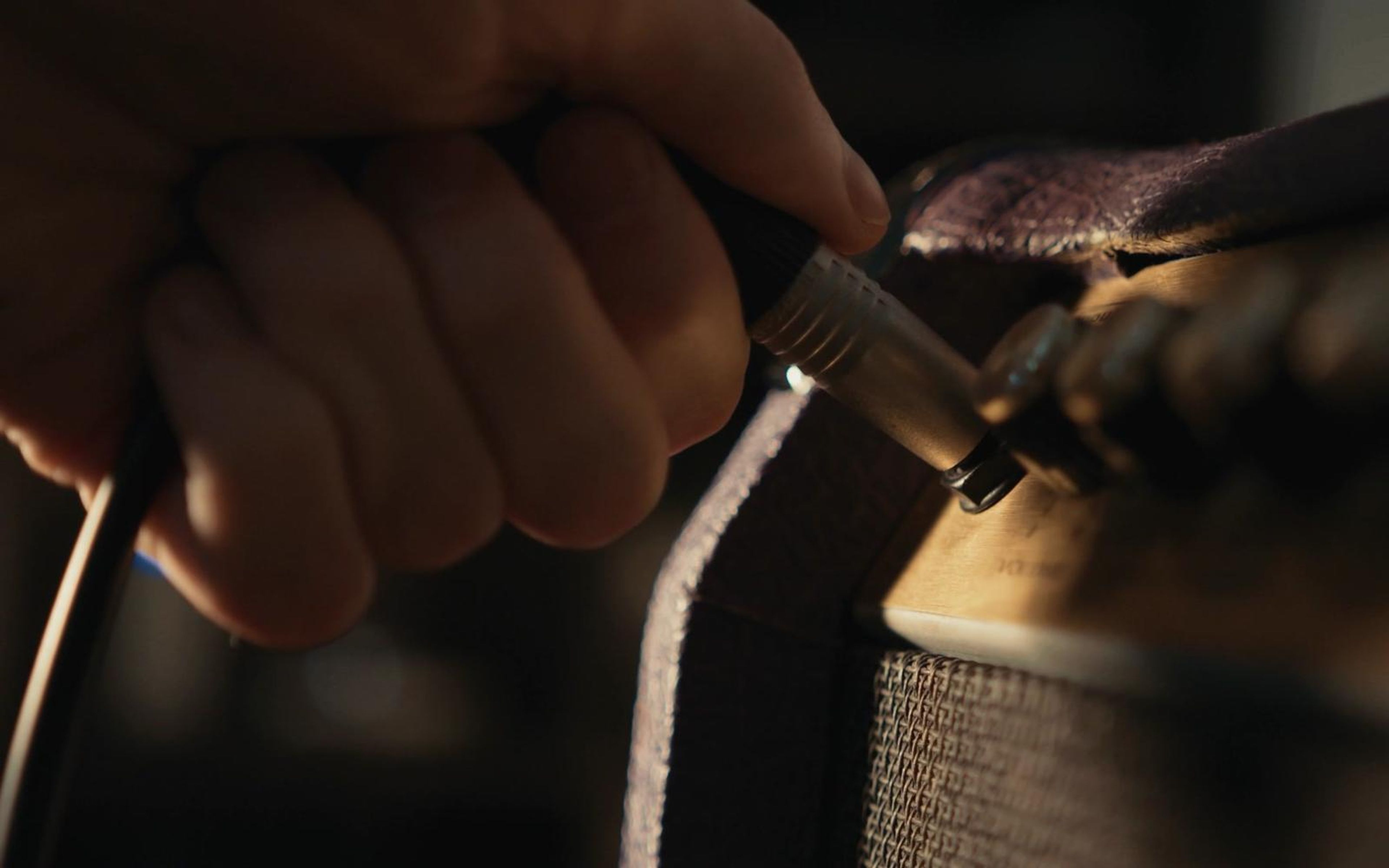 Hand plugging a jack cable into a vintage amplifier with a strap hanging beside it