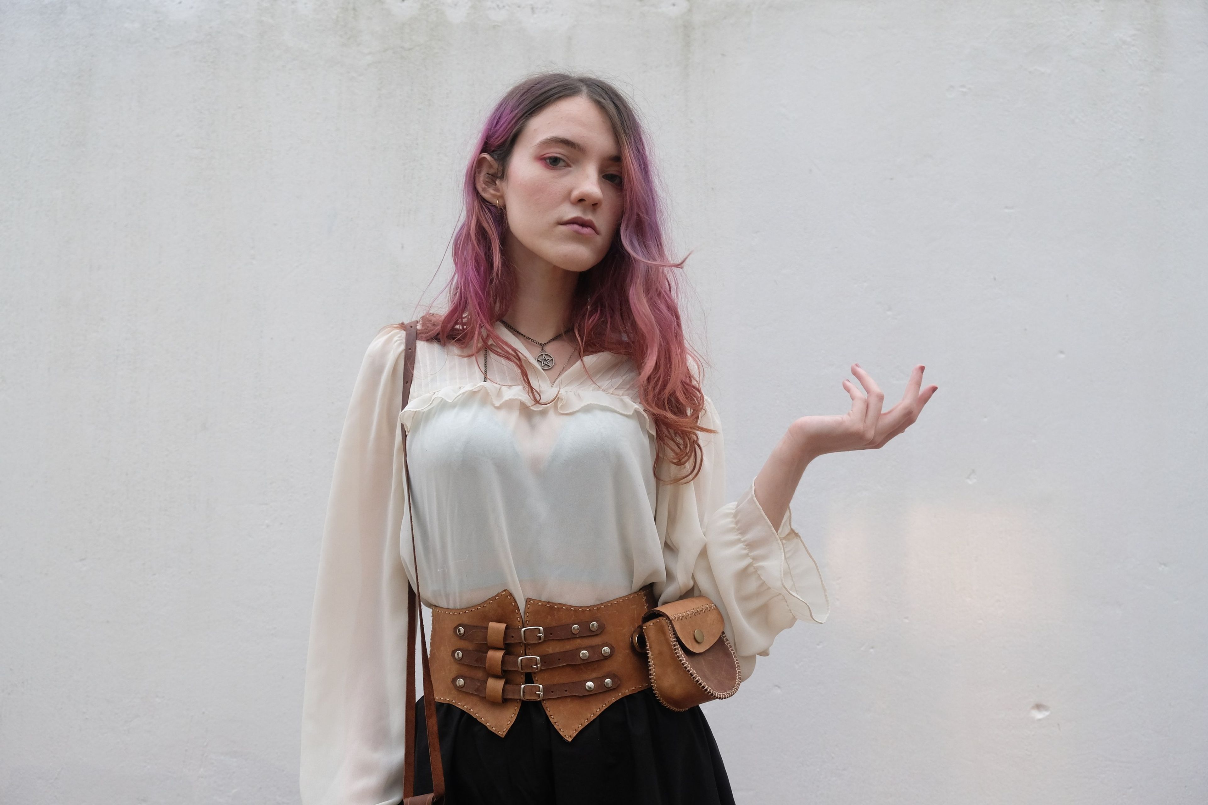 Woman with pink hair holding her hand out, wearing a leather corset belt
