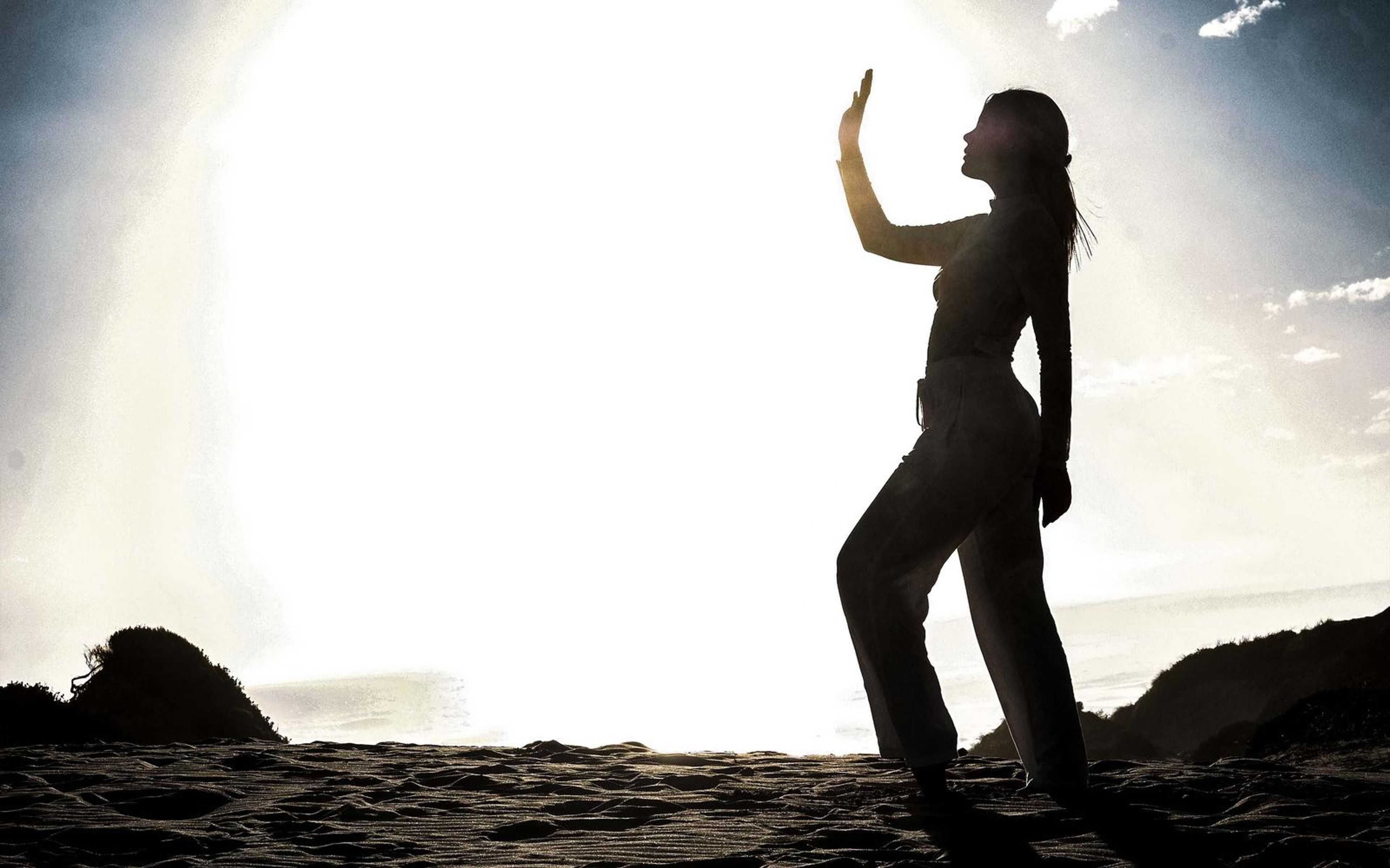 Woman silhouetted on a sand dune with one hand raised toward a bright sky