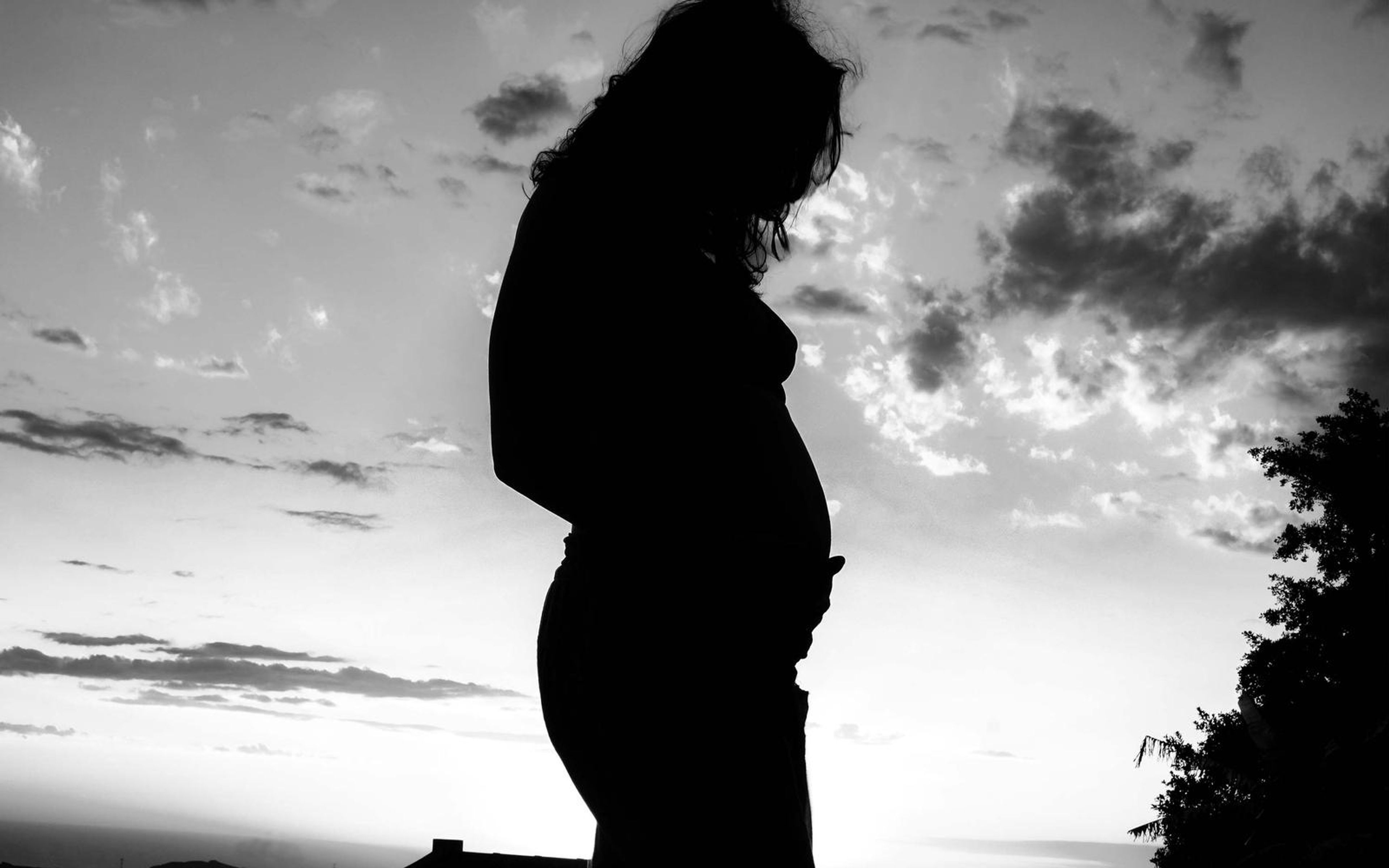 Black and white silhouette of a pregnant woman in profile, tree at the right edge, soft clouds