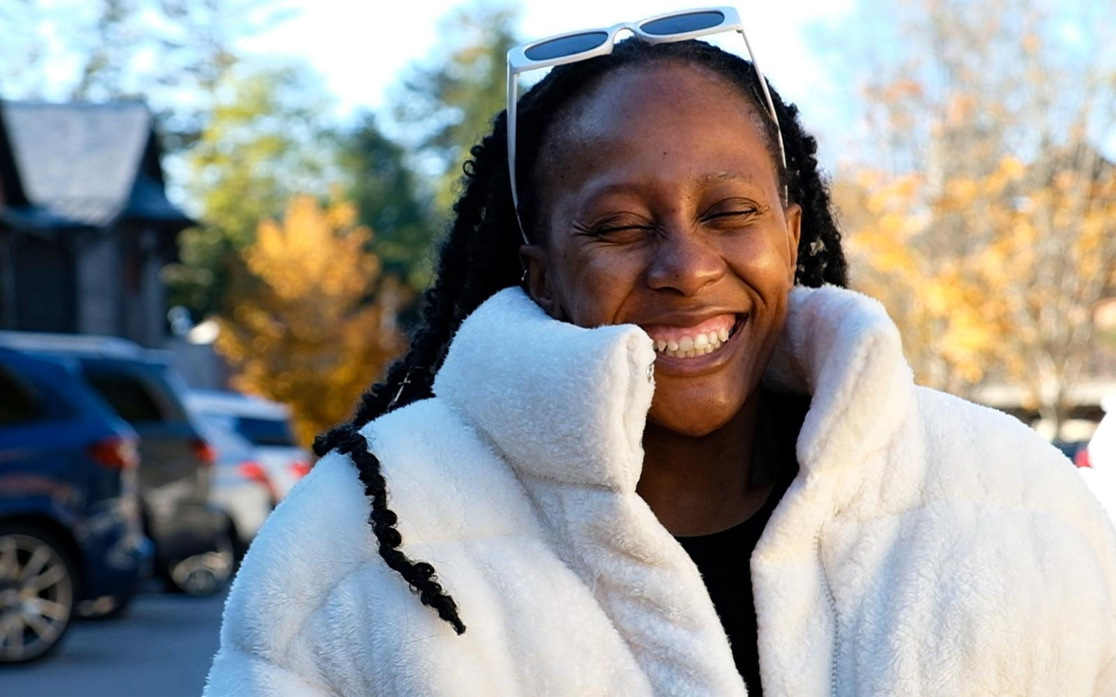 Close image of model laughing with eyes shut, braids over her shoulder, sunglasses on her head, fur collar