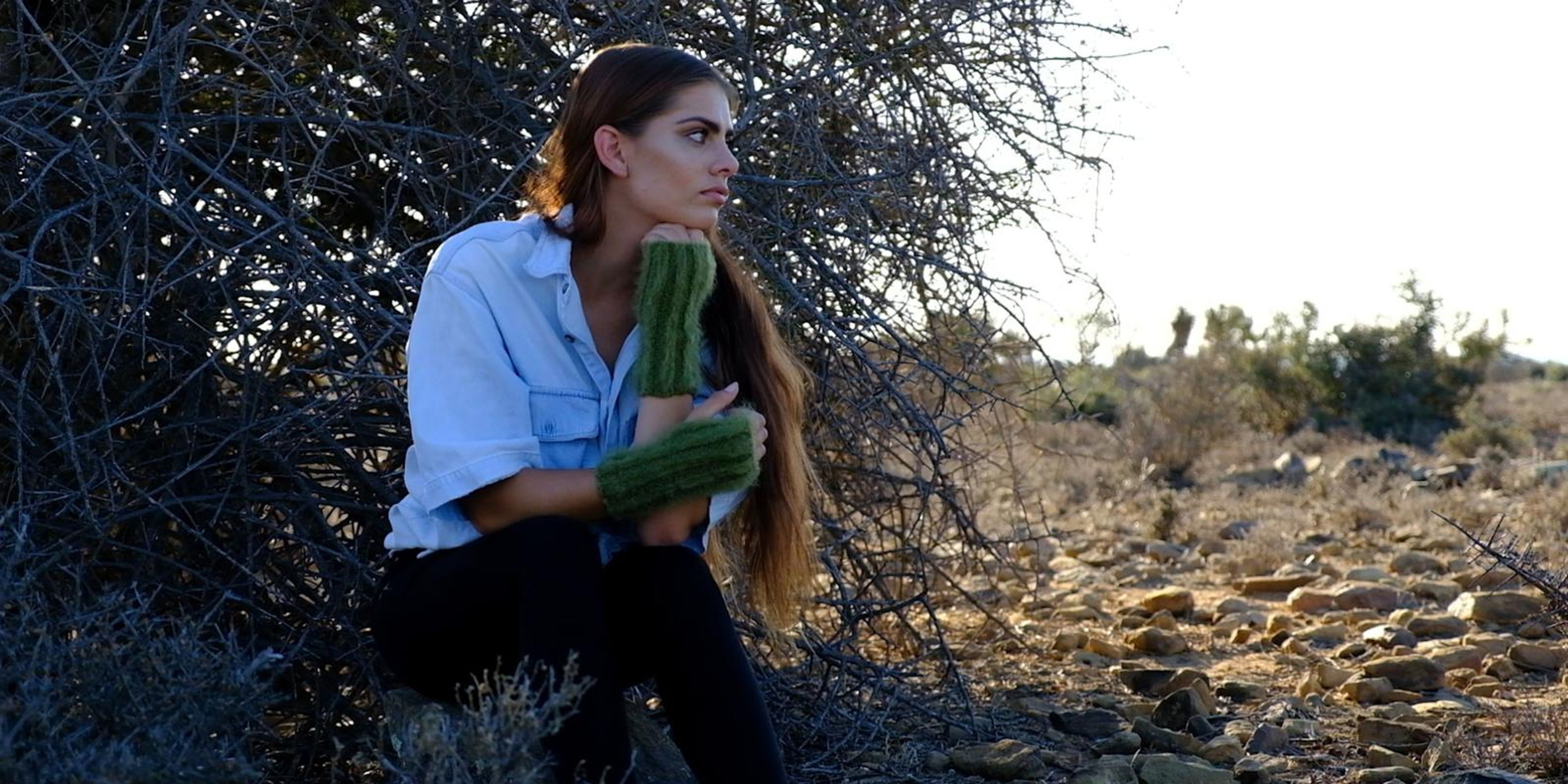 Model seated among thorn bushes holding green mohair fingerless gloves
