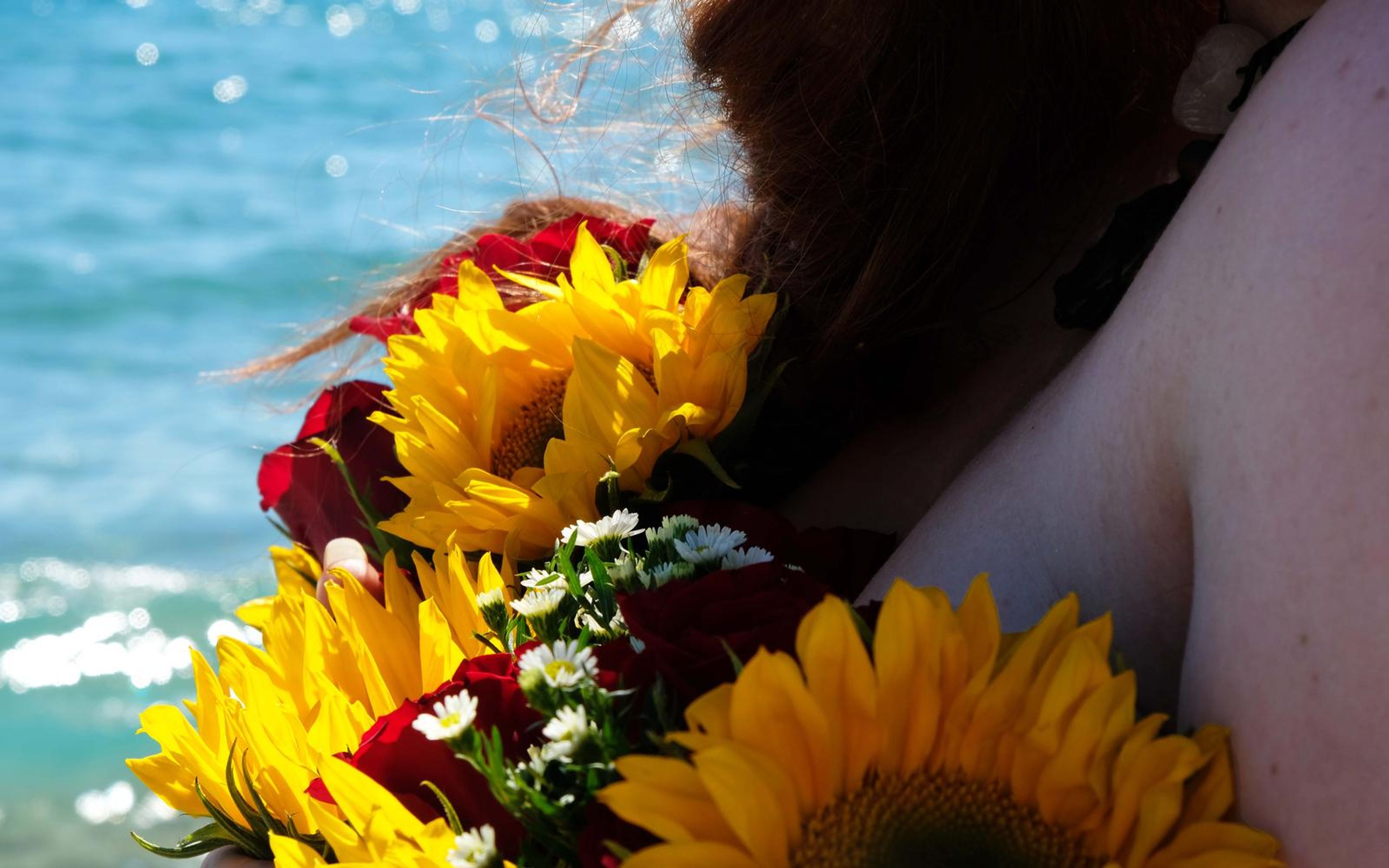 Close crop of a bouquet of sunflowers, red roses and daisies against a bare shoulder, with auburn hair blowing and sparkling sea behind