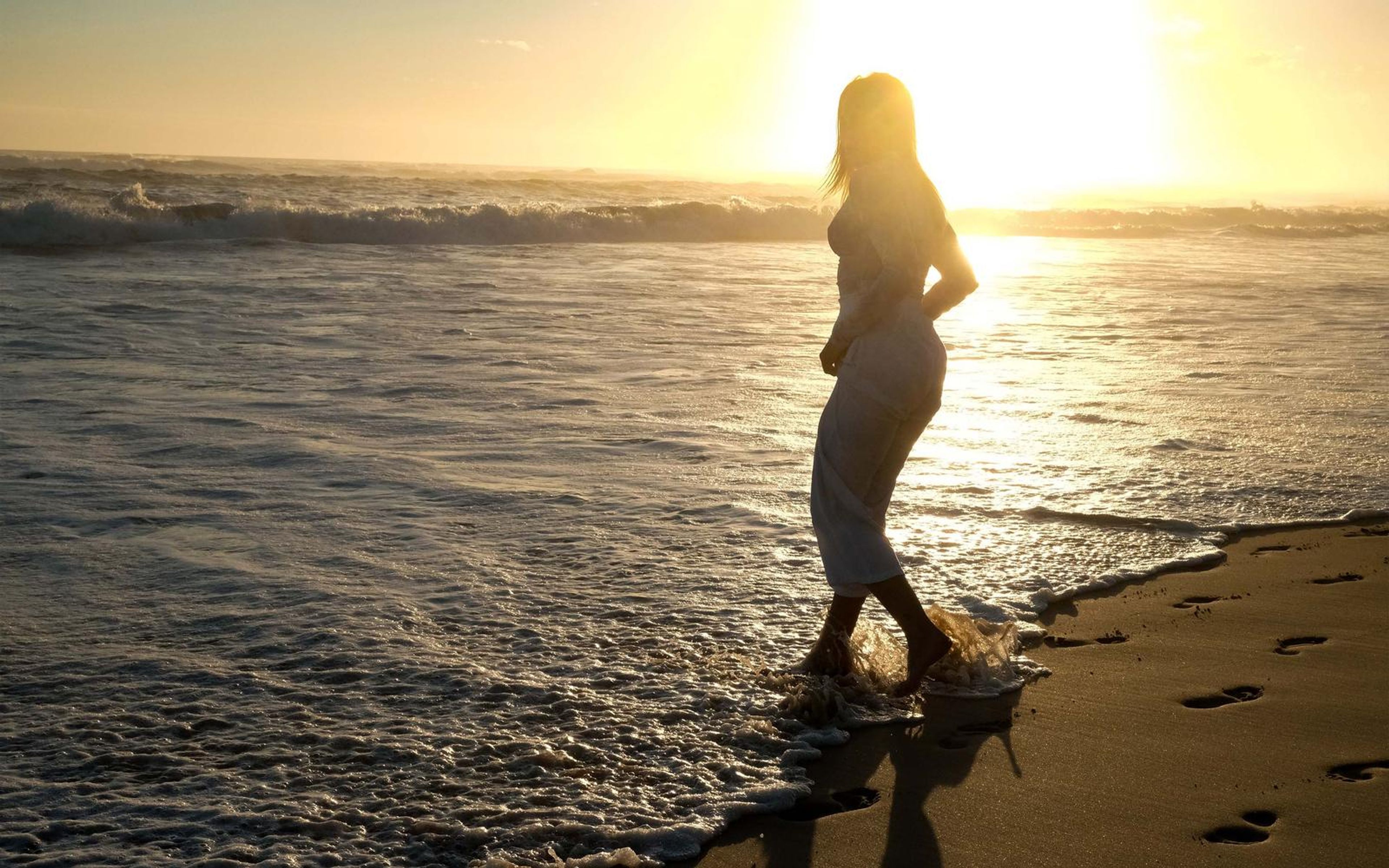 Woman standing in shallow surf as a distant silhouette with footprints on the shore