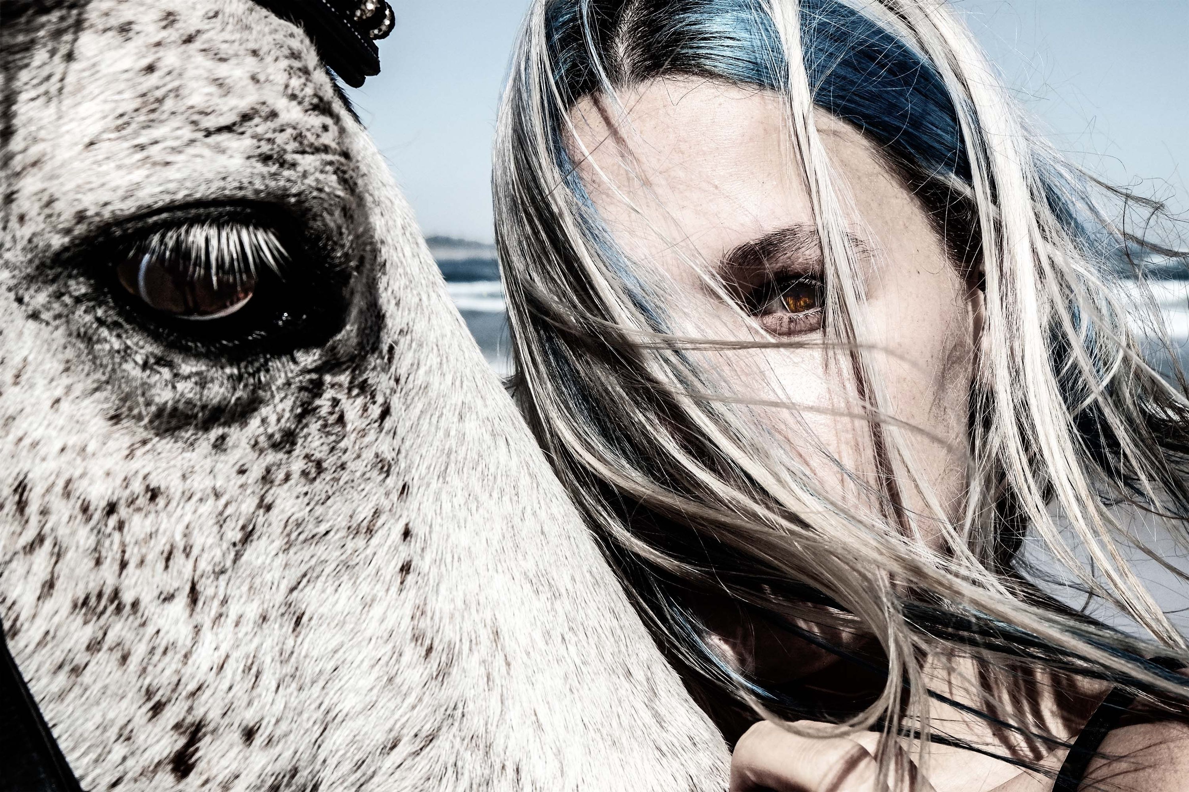 Woman with blonde and blue hair resting her cheek on a white horse's mane, smiling toward camera, beach light