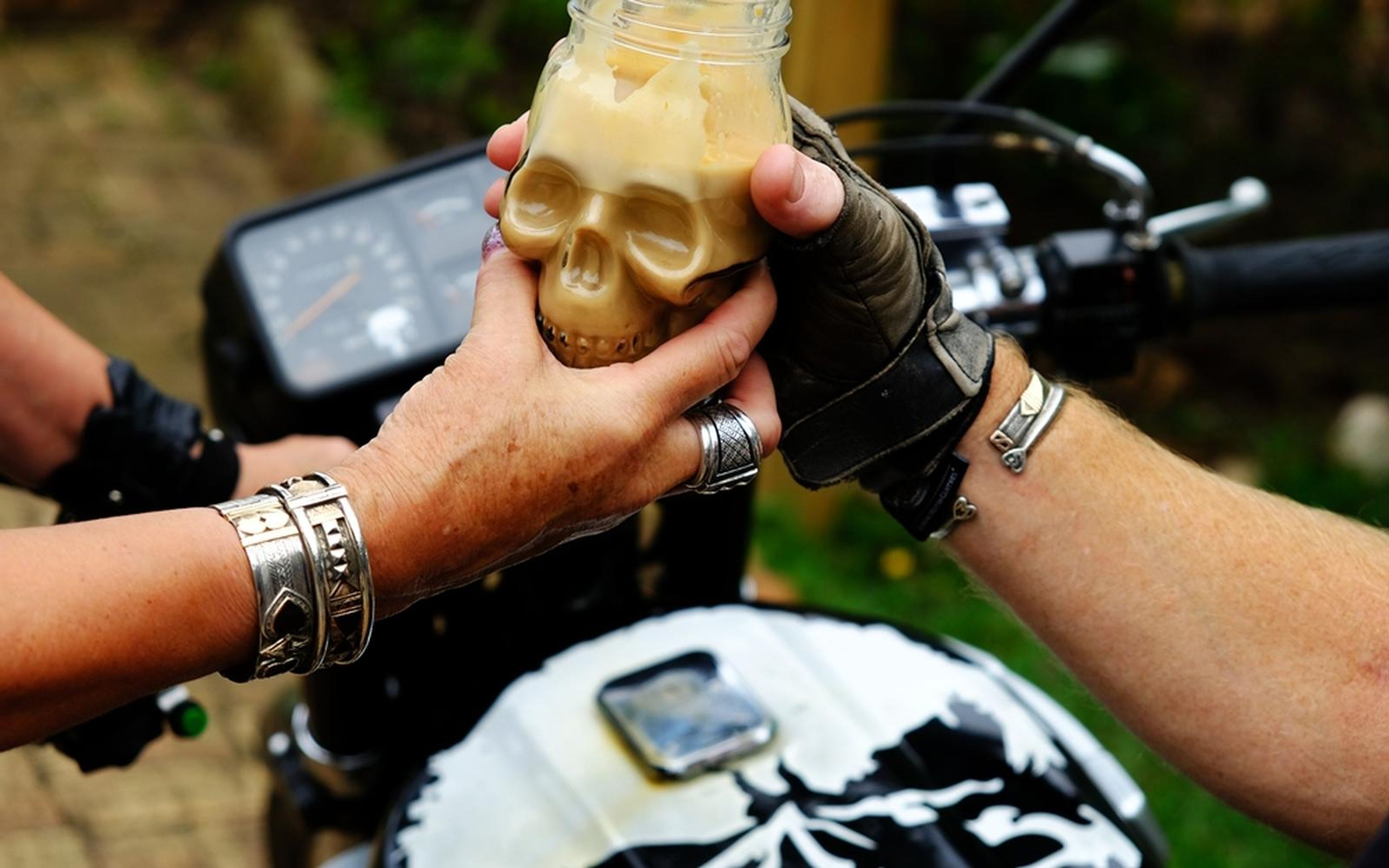 Hands passing a skull-shaped glass jar with a drink, motorbike dashboard behind