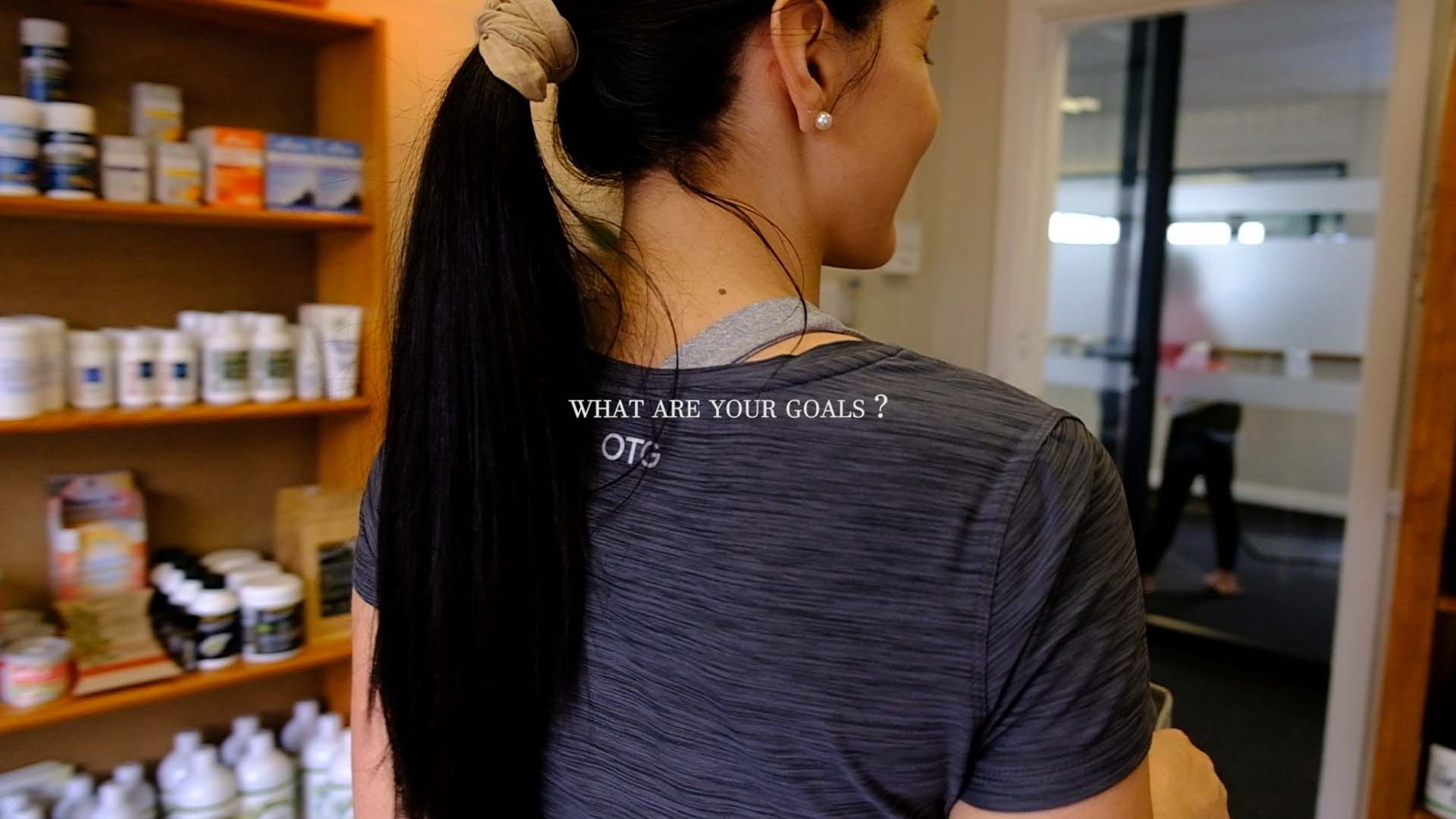 A staff member seen from behind in a grey t-shirt reading 'What are your goals?' in front of shelves of wellness products