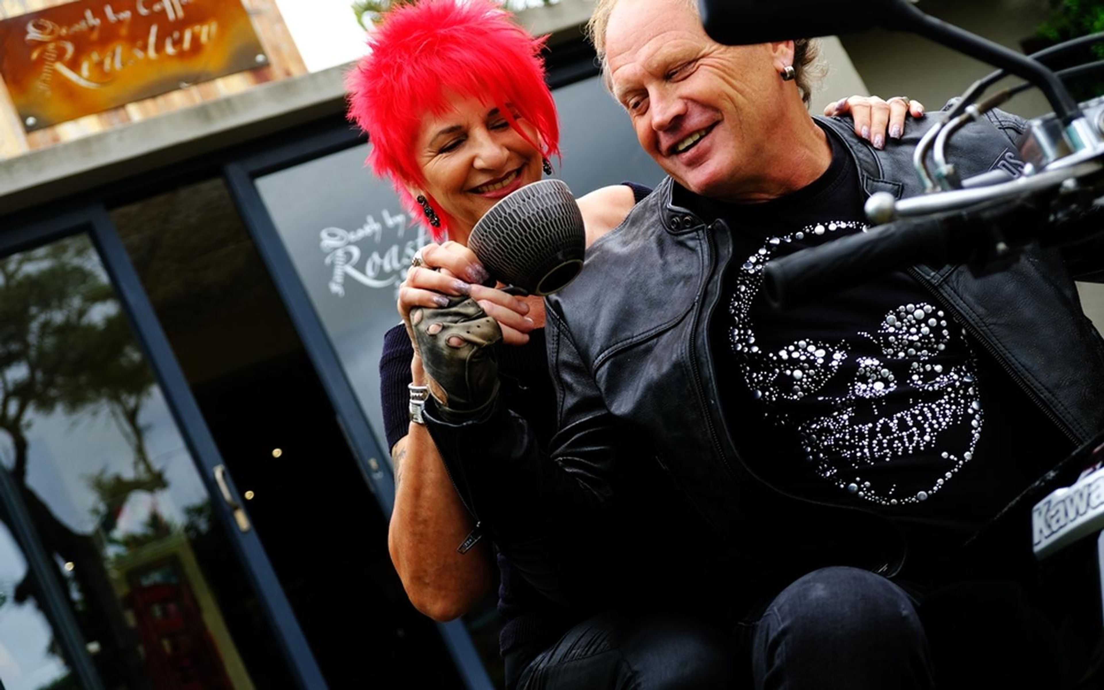 Couple on a parked motorbike laughing together, woman with bright red hair holding a cup, man in leather jacket