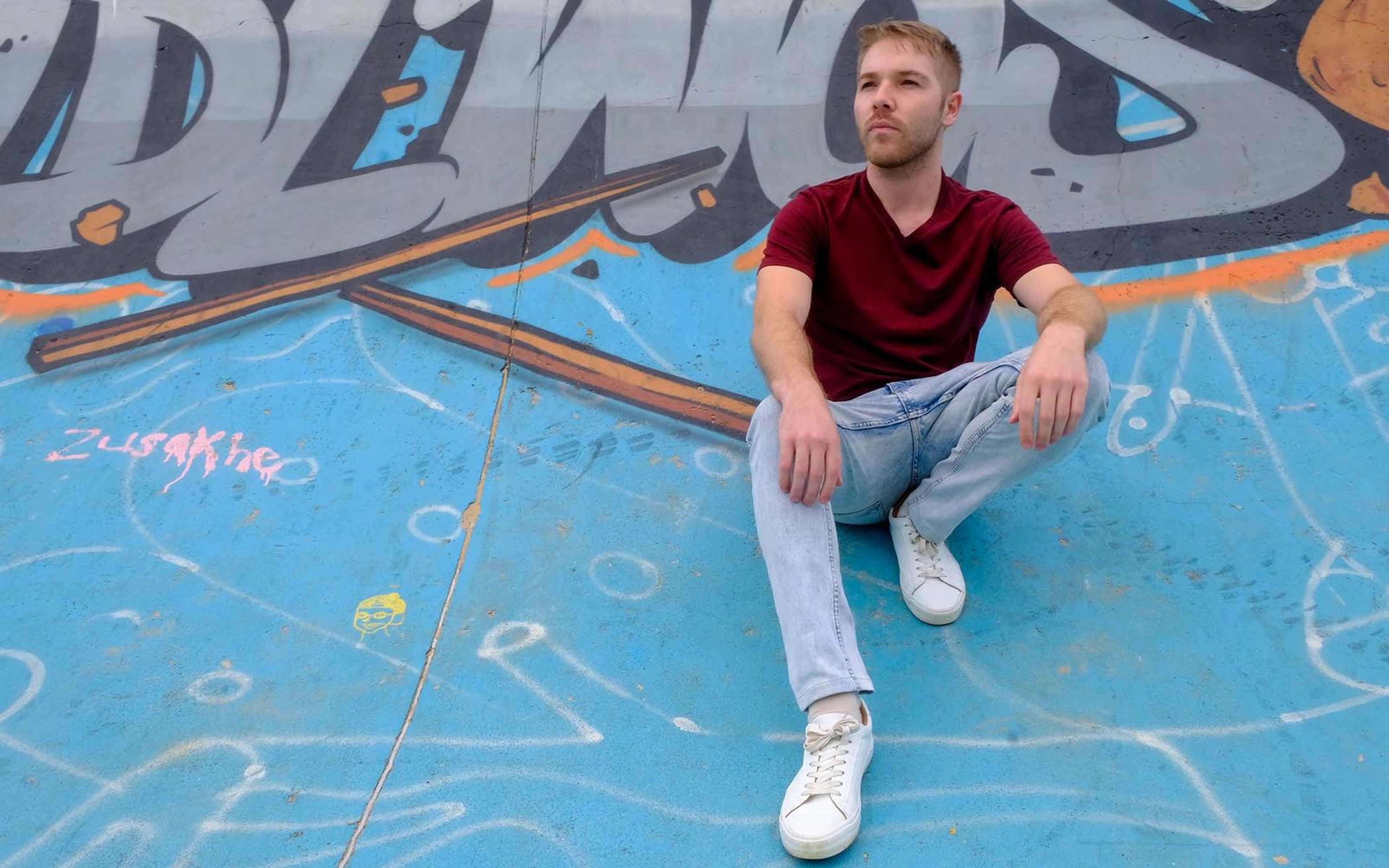 Man in a maroon tee and light jeans seated on a blue painted skate bowl in white sneakers