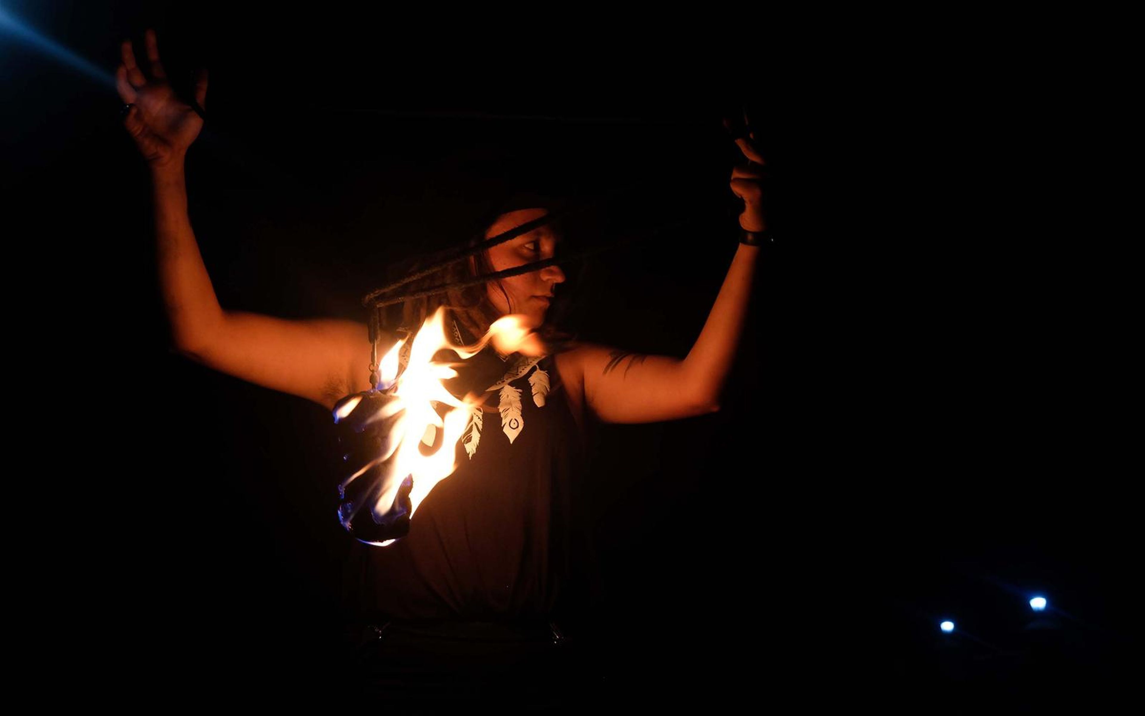 Performer with raised arms in the dark holding a burning prop at the waist, blue lights in the distance