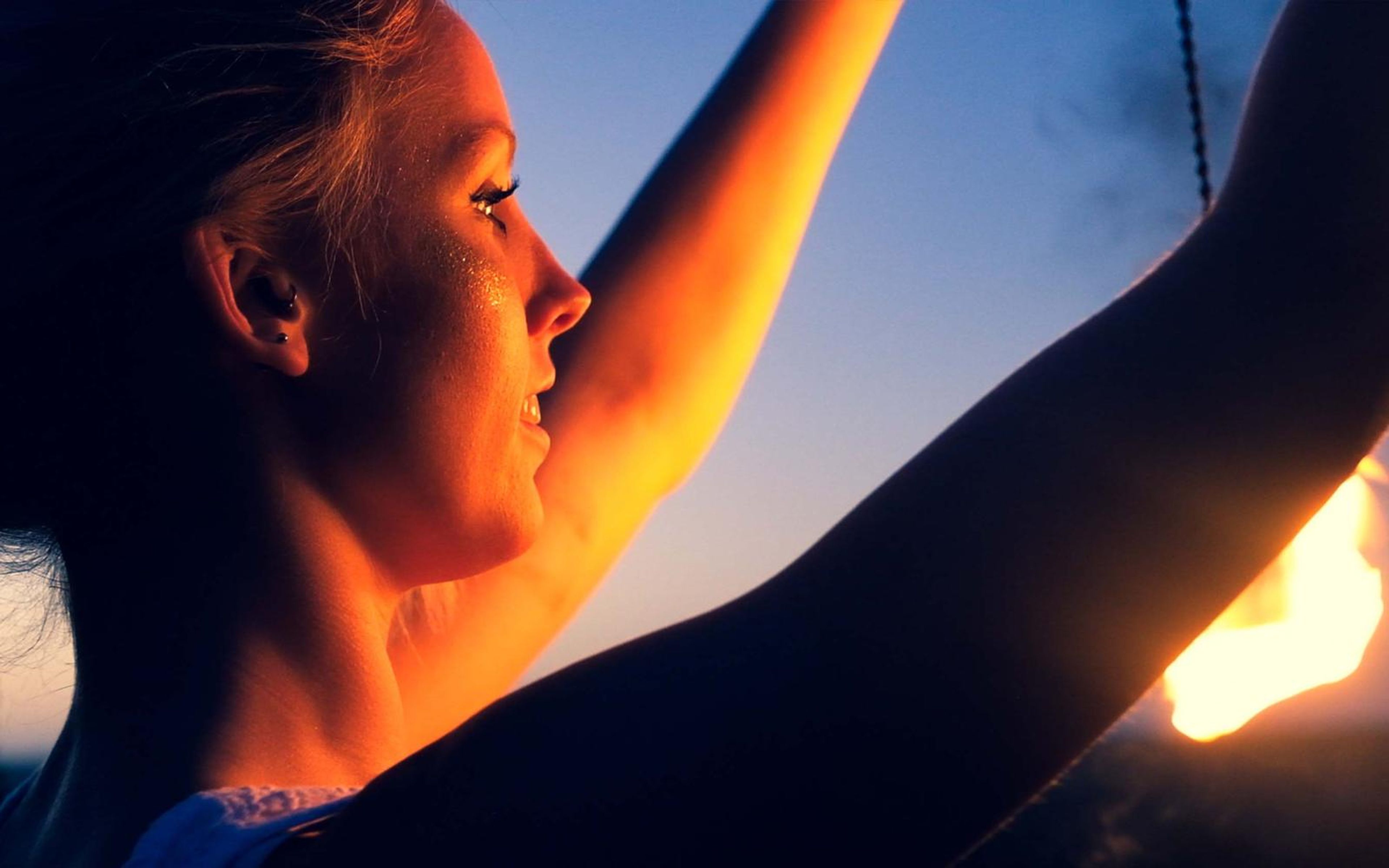 Warm-toned profile close-up of a woman with raised arms, flame at the lower right and a chain hanging behind