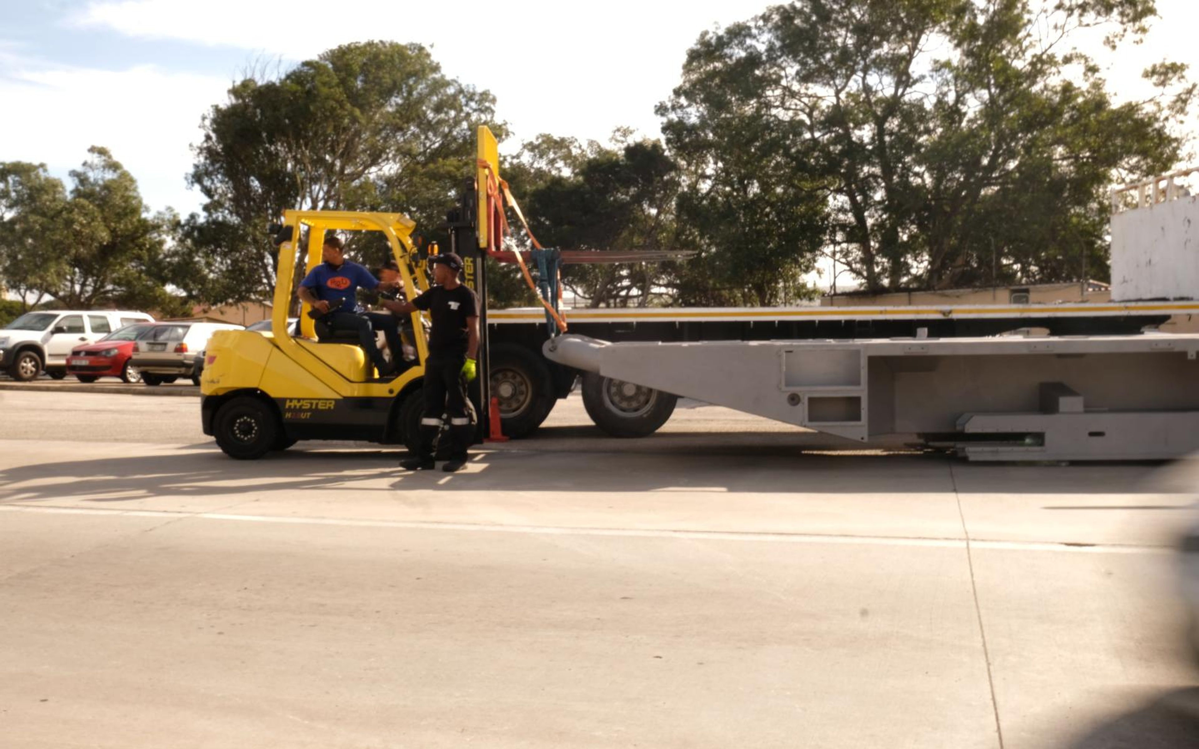 Forklift carrying a grey primed steel chassis section on a flatbed in the yard