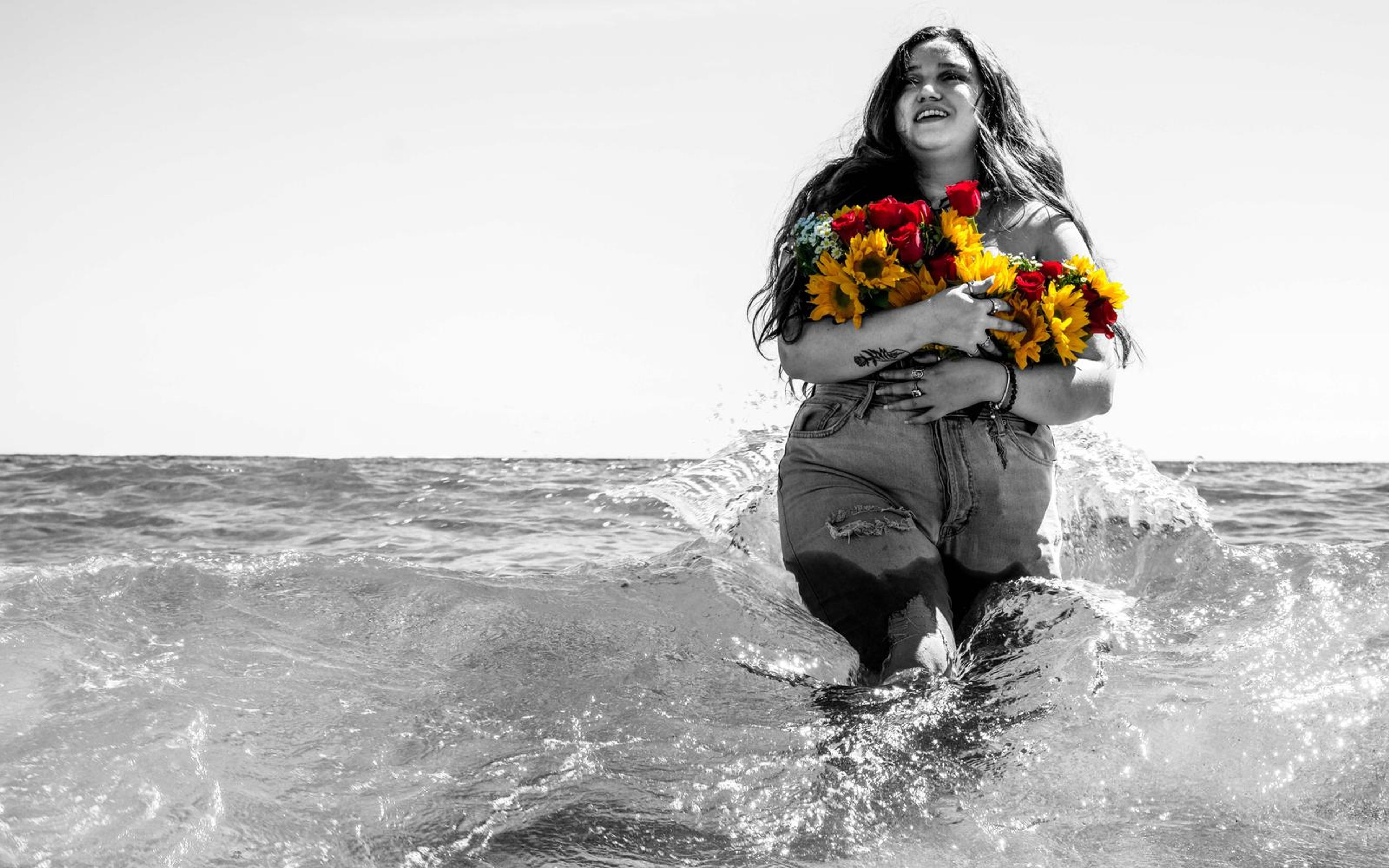Black and white image of model smiling as water splashes around her, flowers in her arms