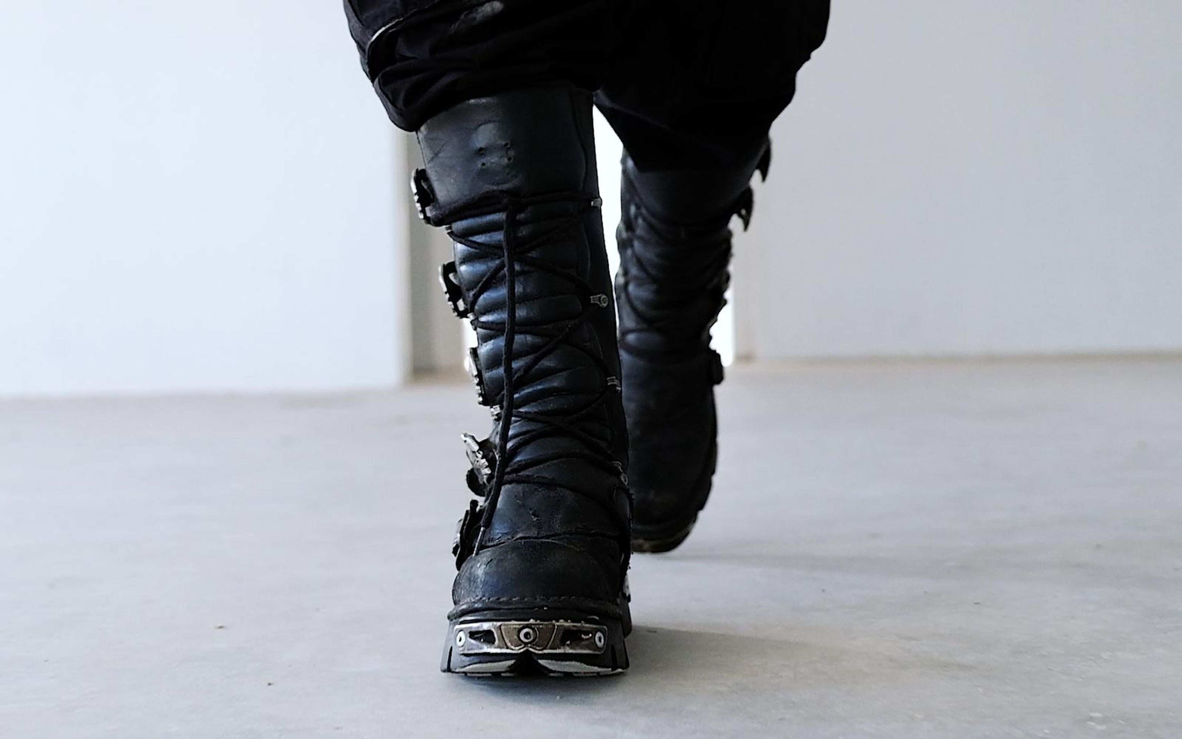 Low view of black laced boots mid-stride with a metal toe plate, blurred white room behind