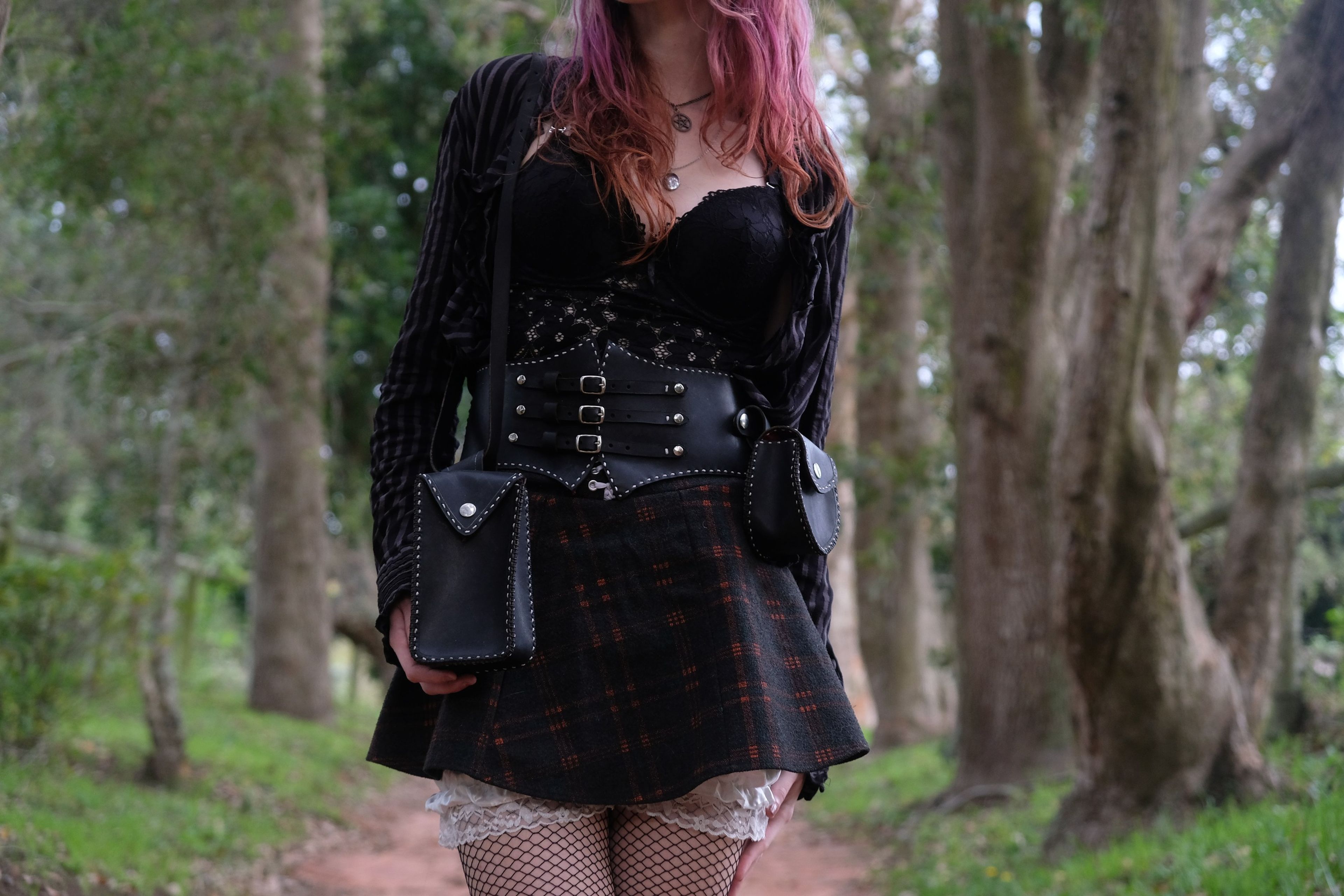 Woman in a black top and corset belt with a bag at her hip, among tall trees