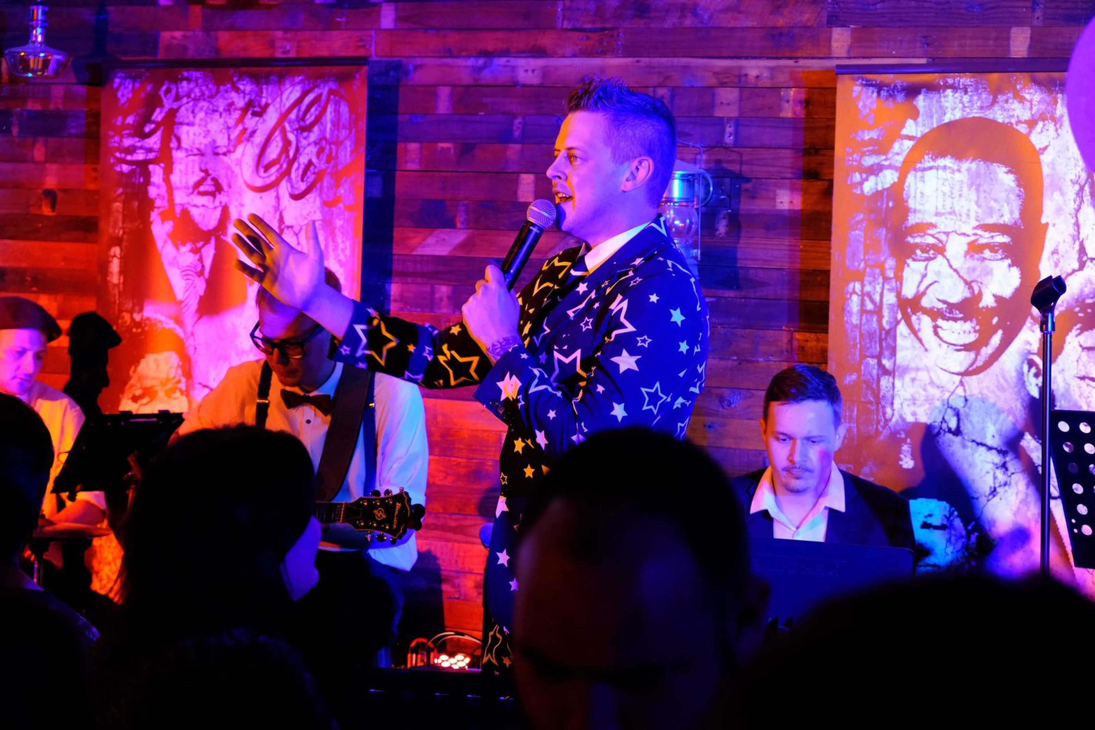 Singer in a star-patterned suit gesturing in front of a mural of a smiling face