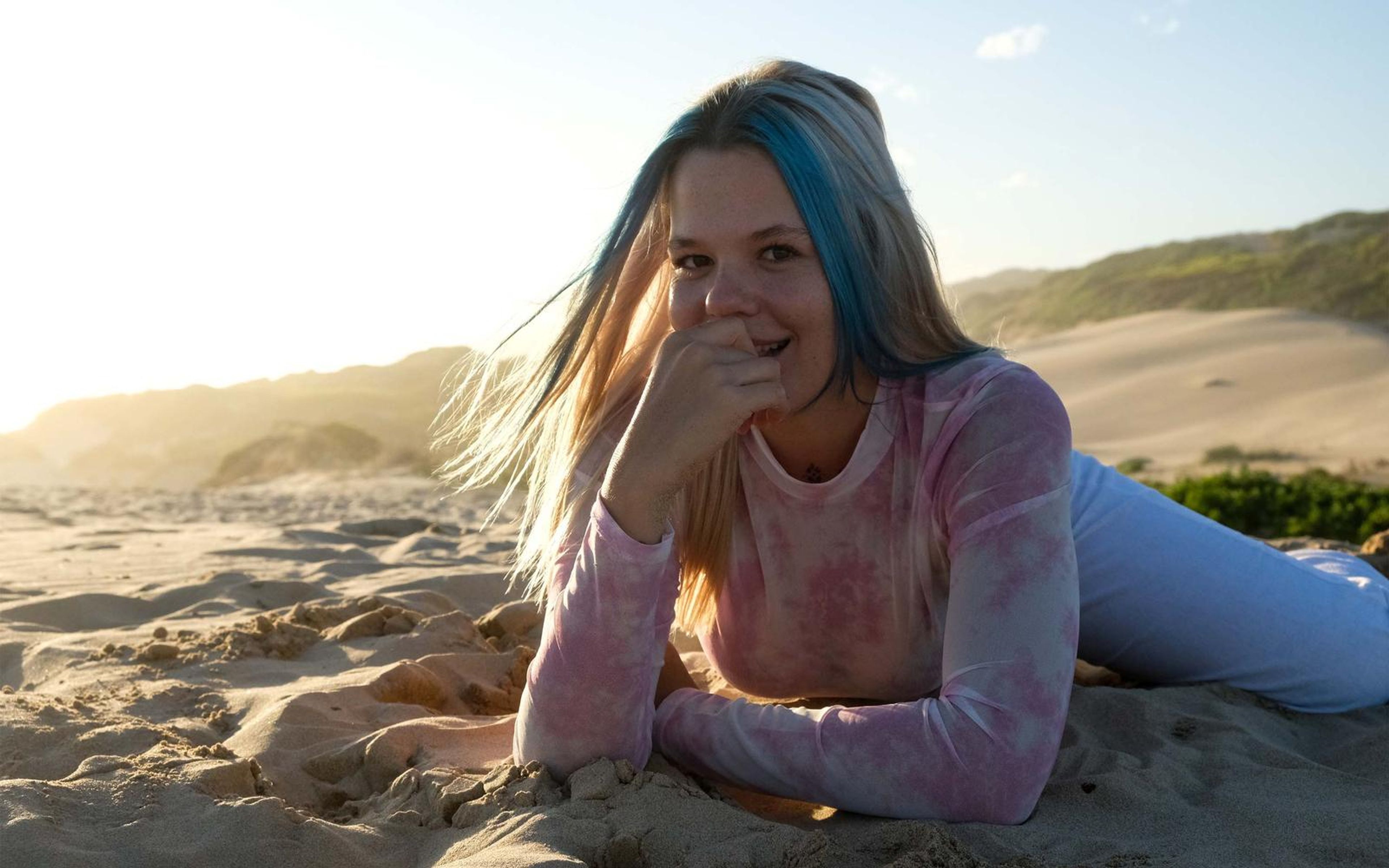 Woman lying on sand laughing with her hand covering her mouth, dunes and warm haze behind