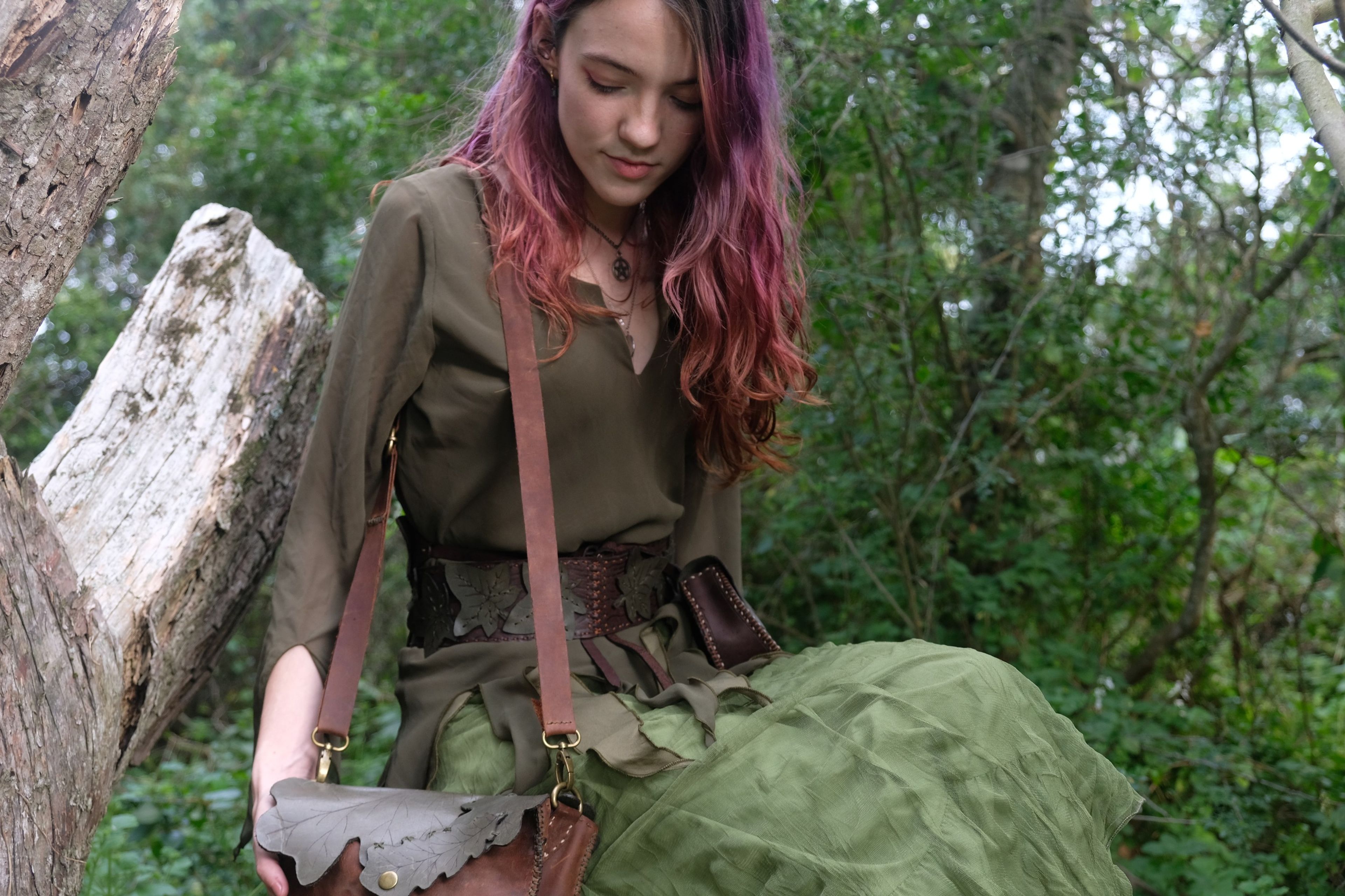 Woman with pink hair holding a leather bag, seated by a fallen log