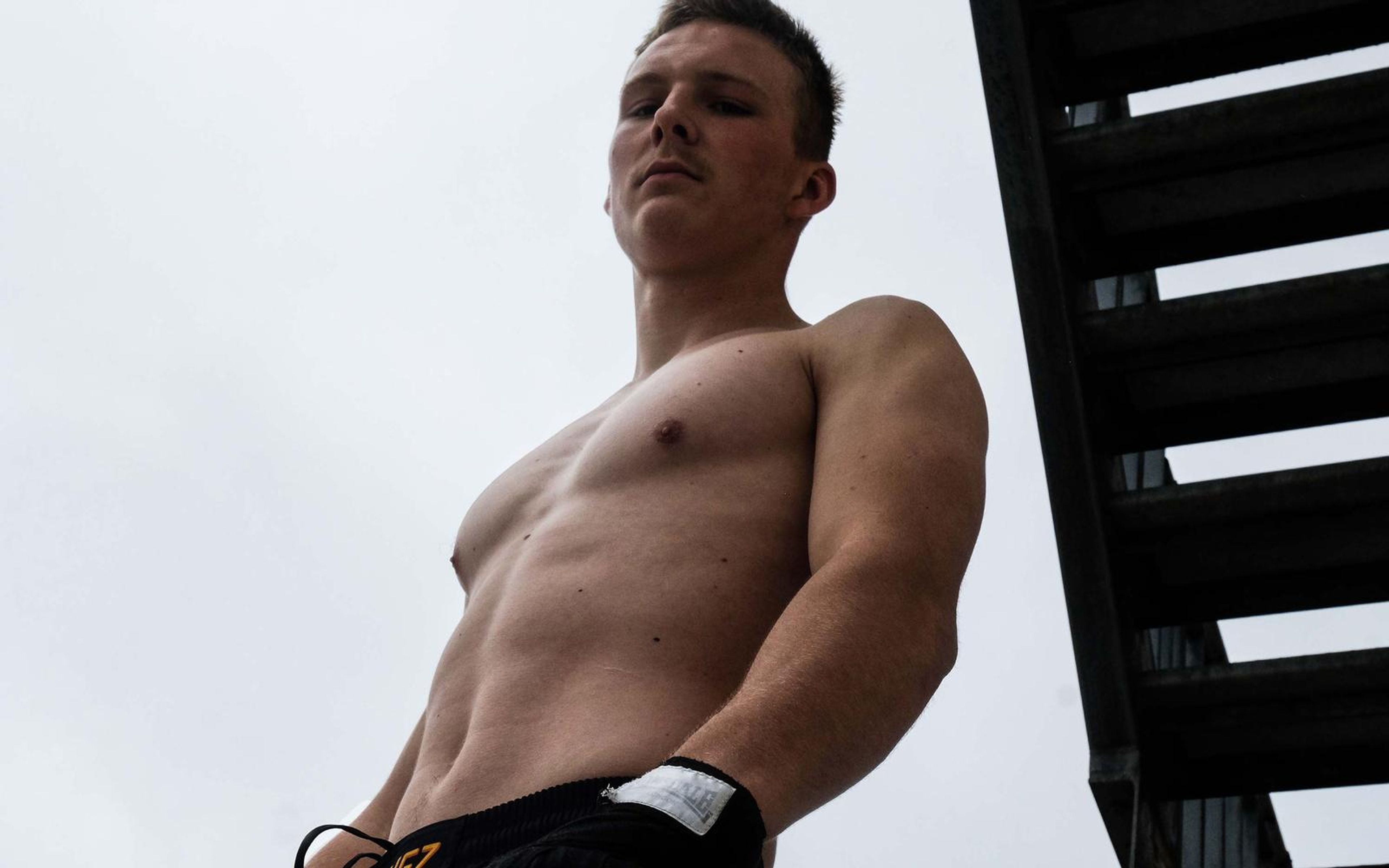 Fighter in black shorts seen from a low angle against a white sky beside a steel fire stair, bare torso