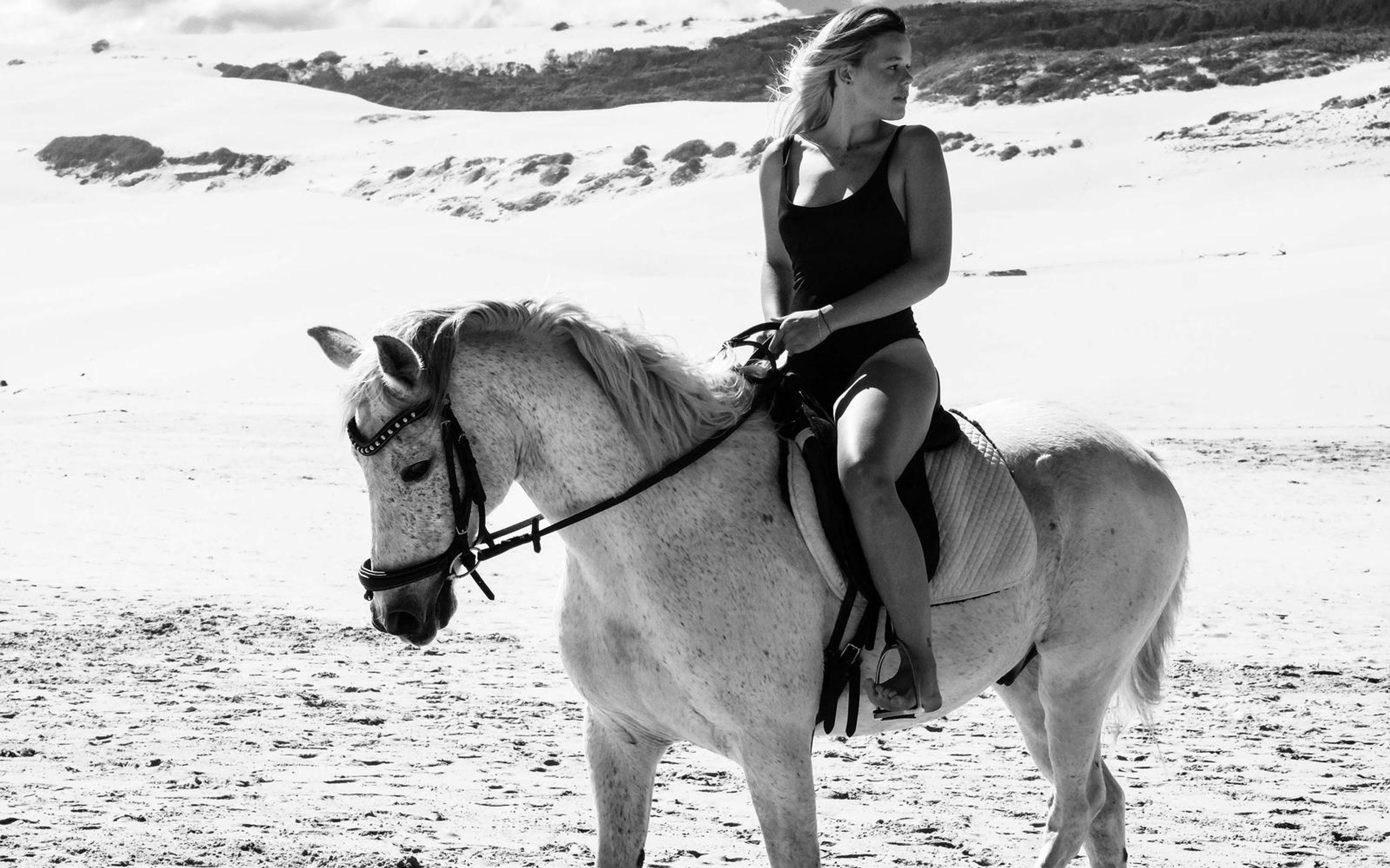 Black and white image of a woman riding a white horse across open sand with dune ridges behind