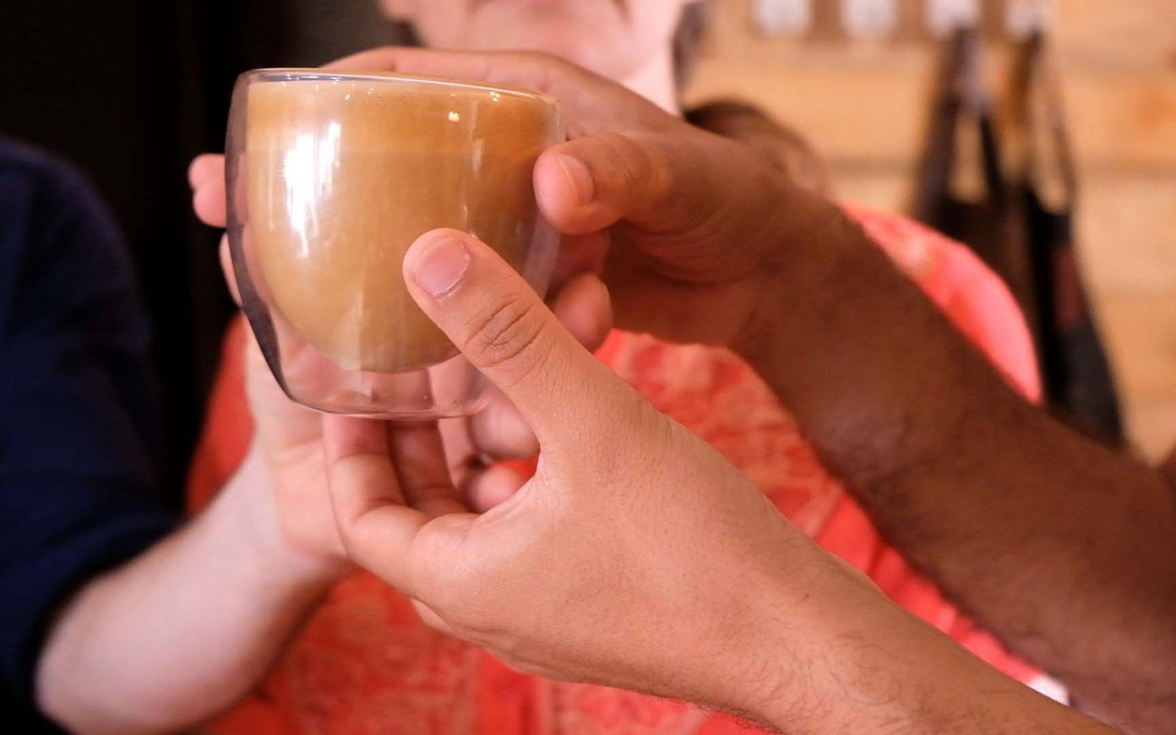 Hands passing a small glass cup of milky coffee to another pair of hands in warm cafe light