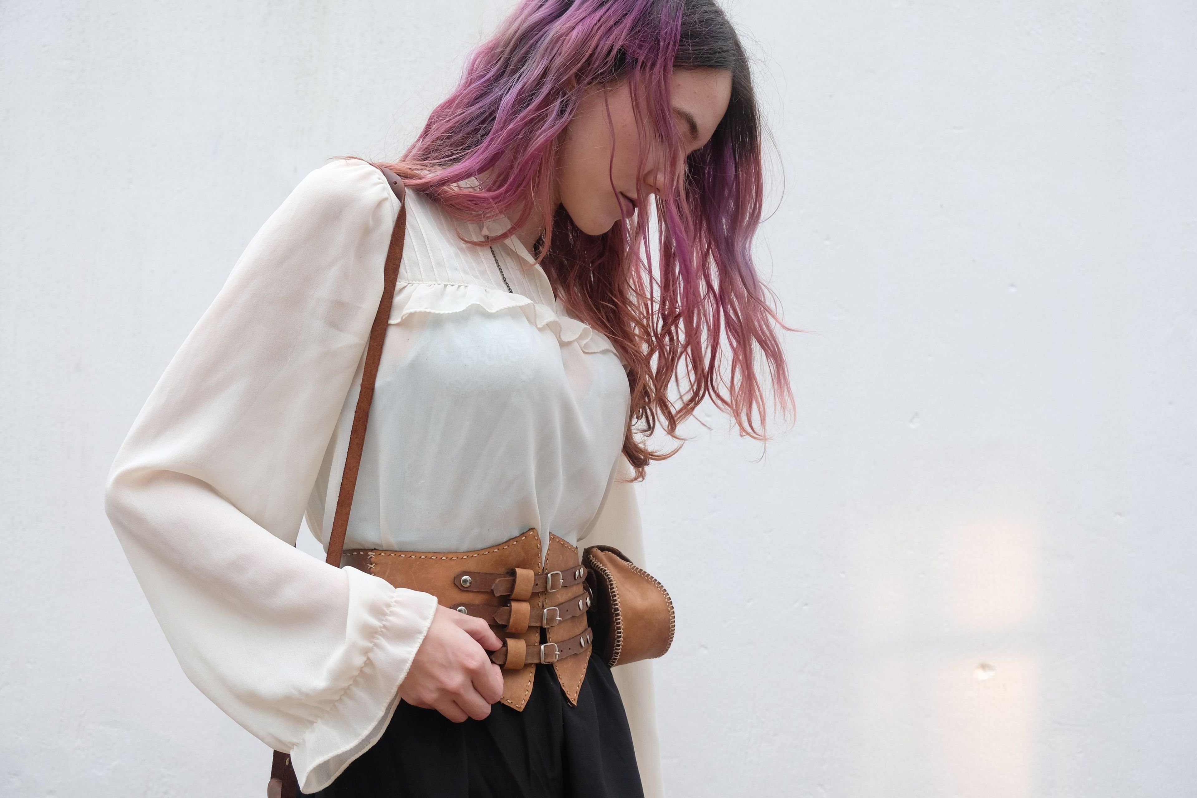 Woman with pink hair looking down, wearing a leather corset belt, cream blouse and shoulder bag