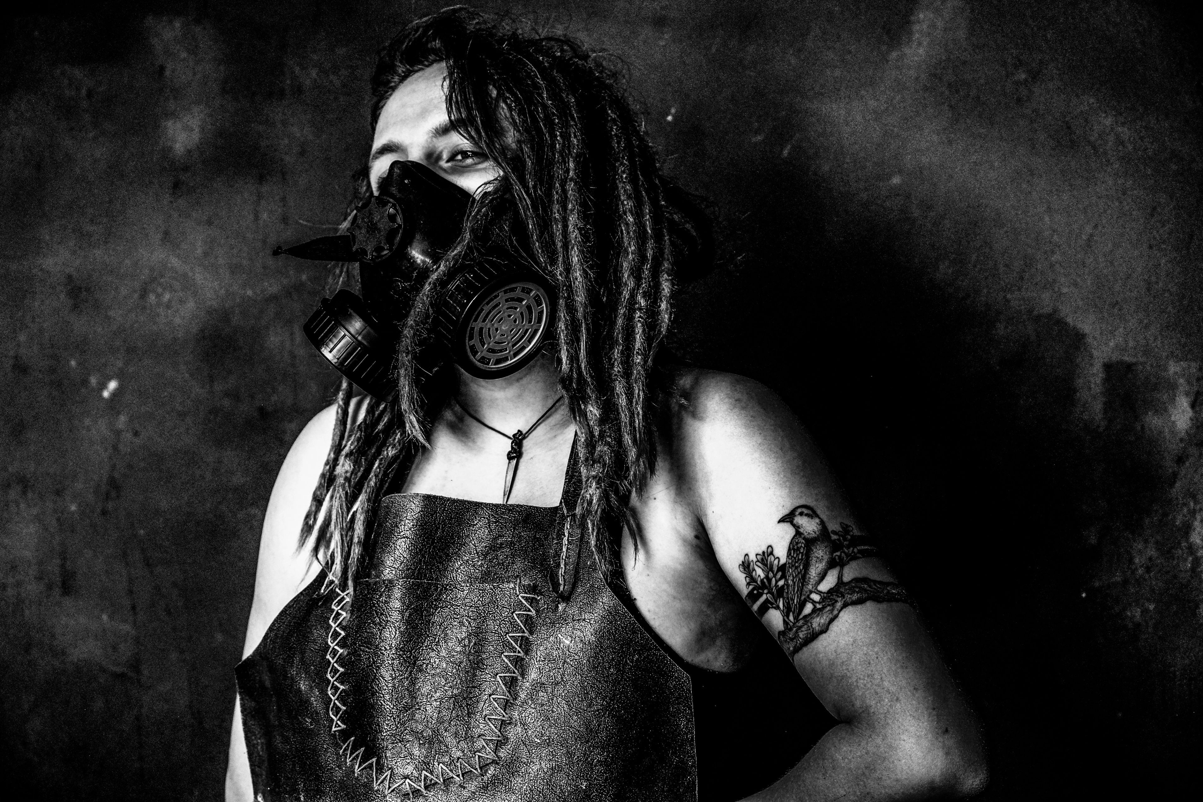 Black and white three-quarter portrait, subject in a leather apron with a gas mask resting below the chin, bird tattoo on the shoulder