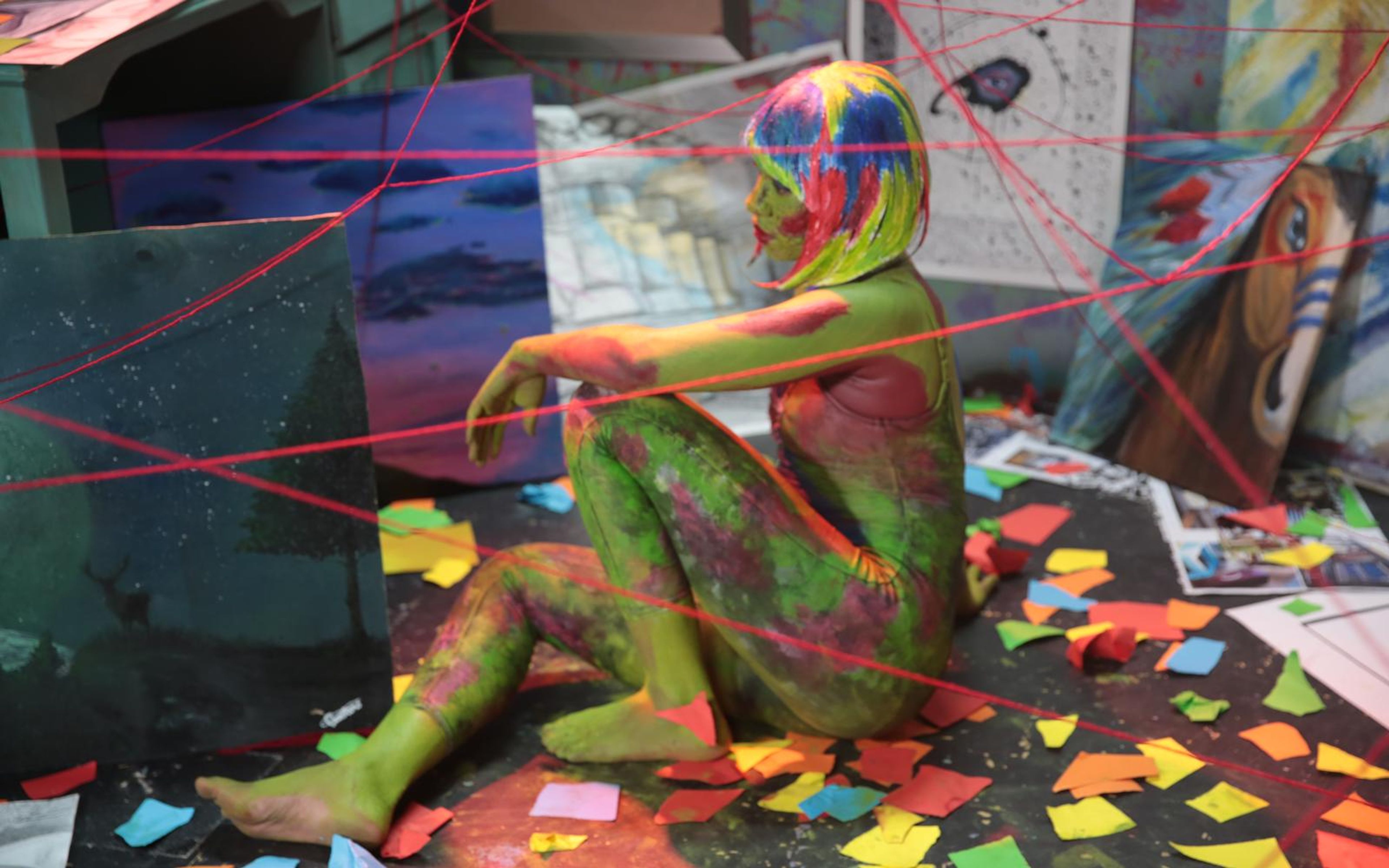 Painted figure seated among paintings and red string in the art room set, shot at an angle