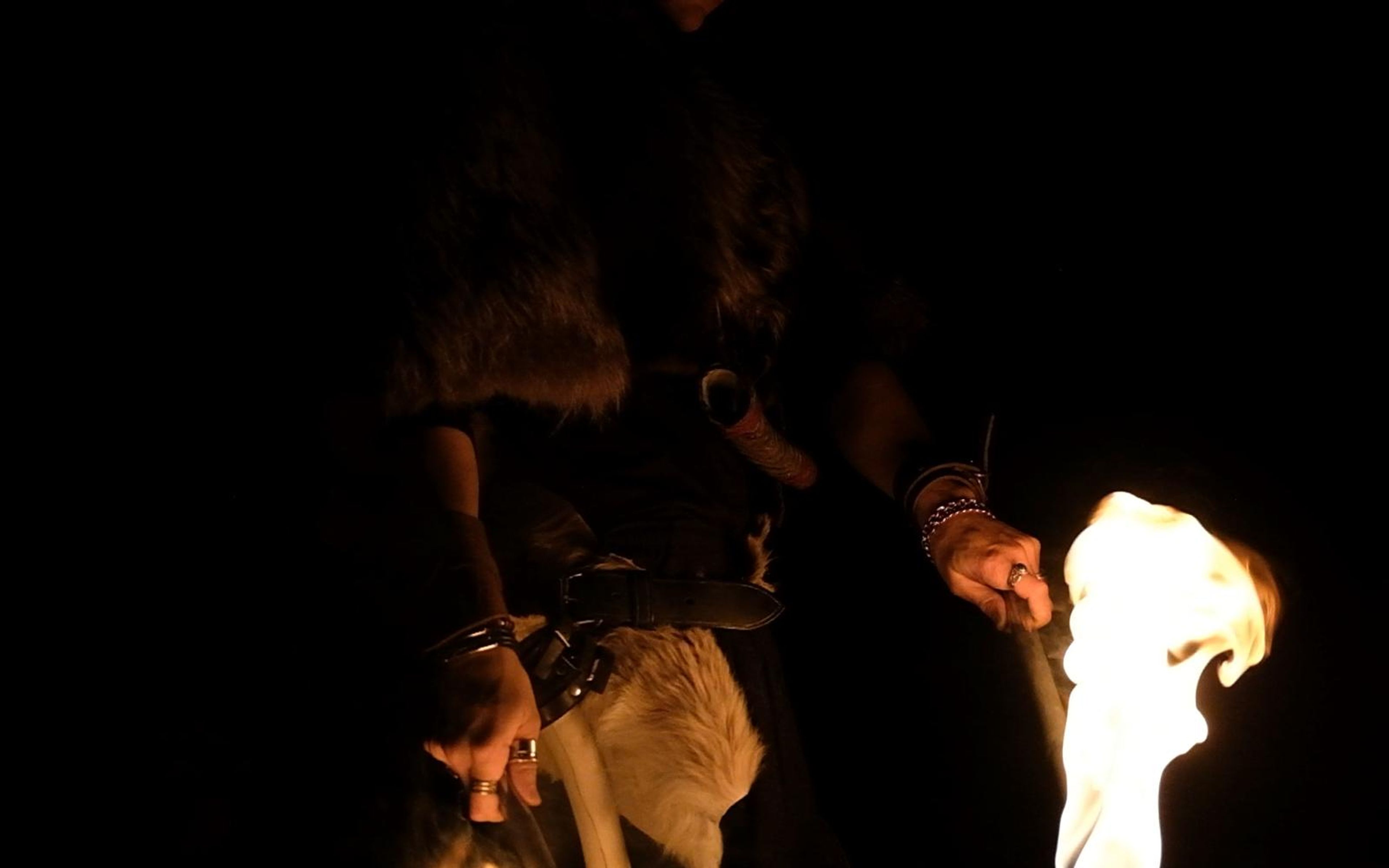 Close view of a large torch flame at right beside a fur cape and leather belt with ring bracelets