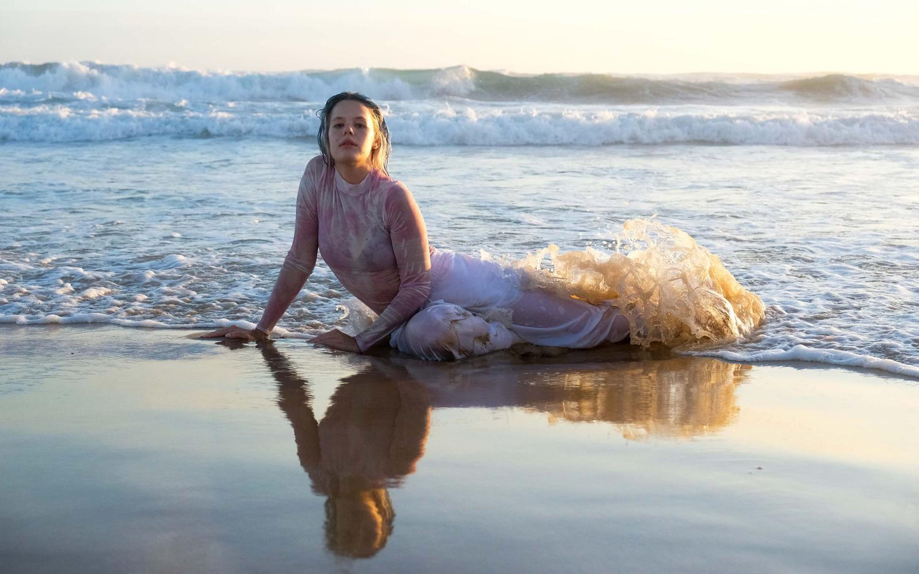 Woman reclining in shallow water at dusk with trousers fanning out in a splash, reflection on the wet sand