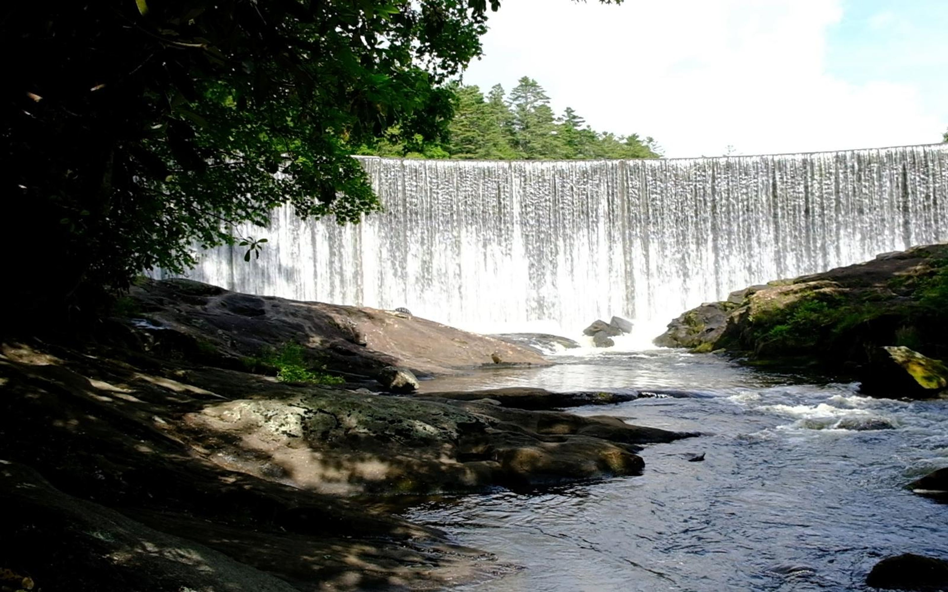 Waterfall in the distance seen from the shaded riverbank, framed by dark branches and flowing water