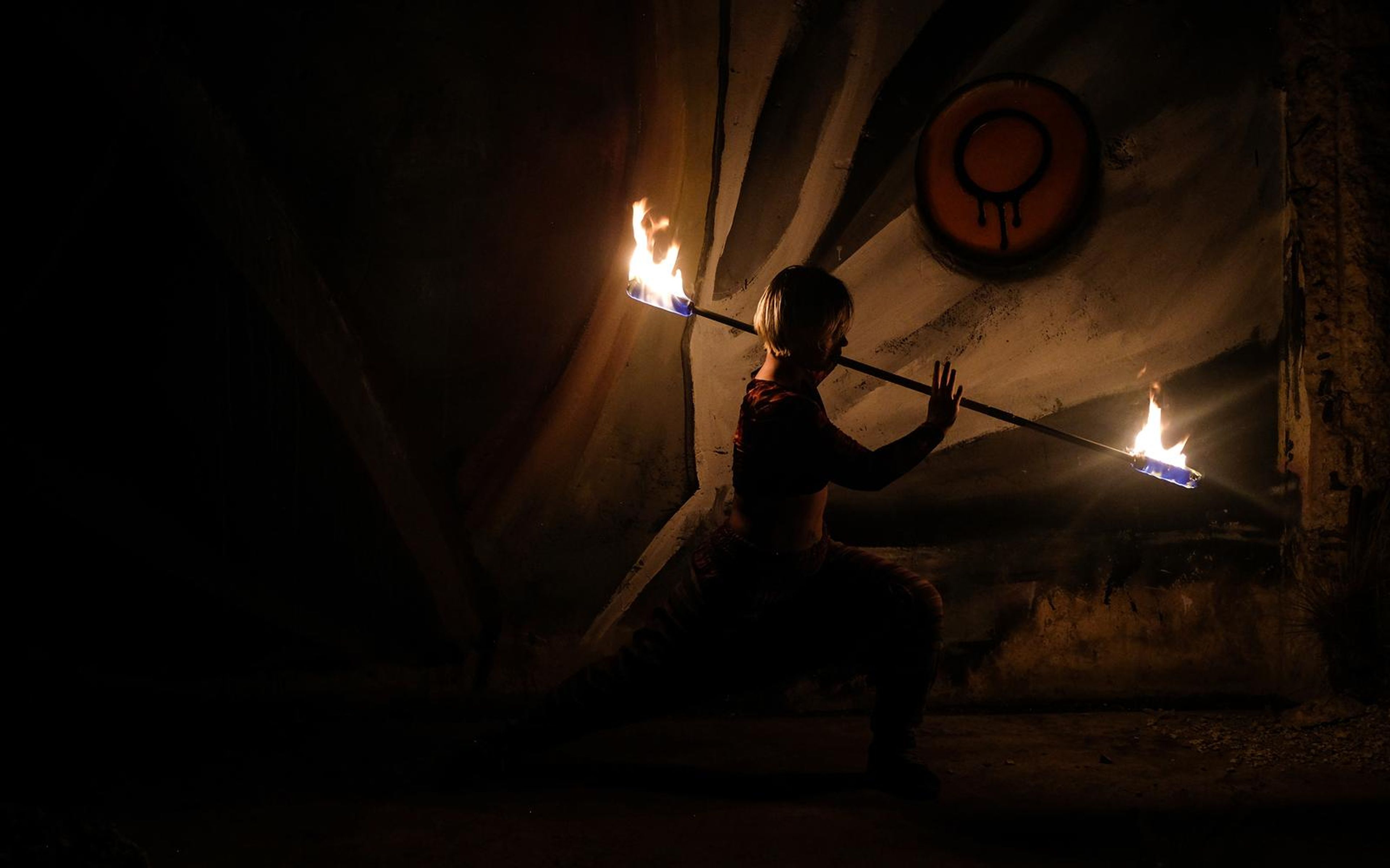 Wide dark shot of a performer crouching with a burning staff beside a painted concrete wall