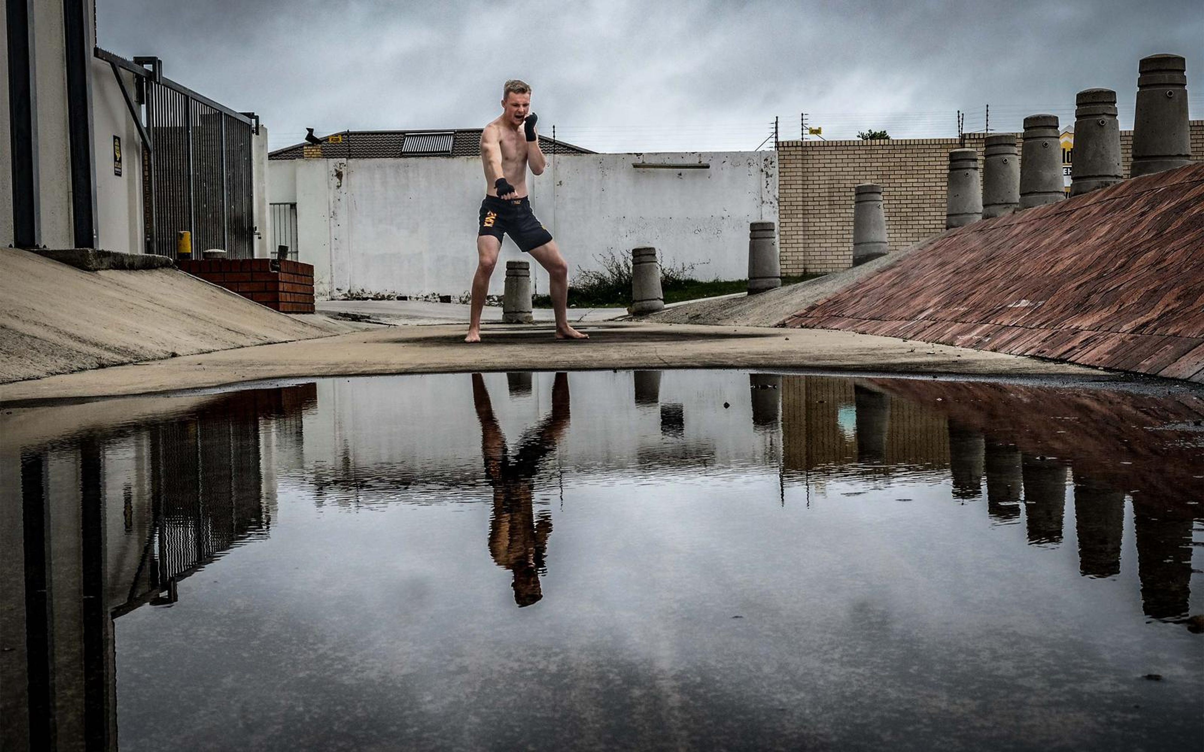 Man standing with fists up and legs apart, mirrored in rain water, heavy sky