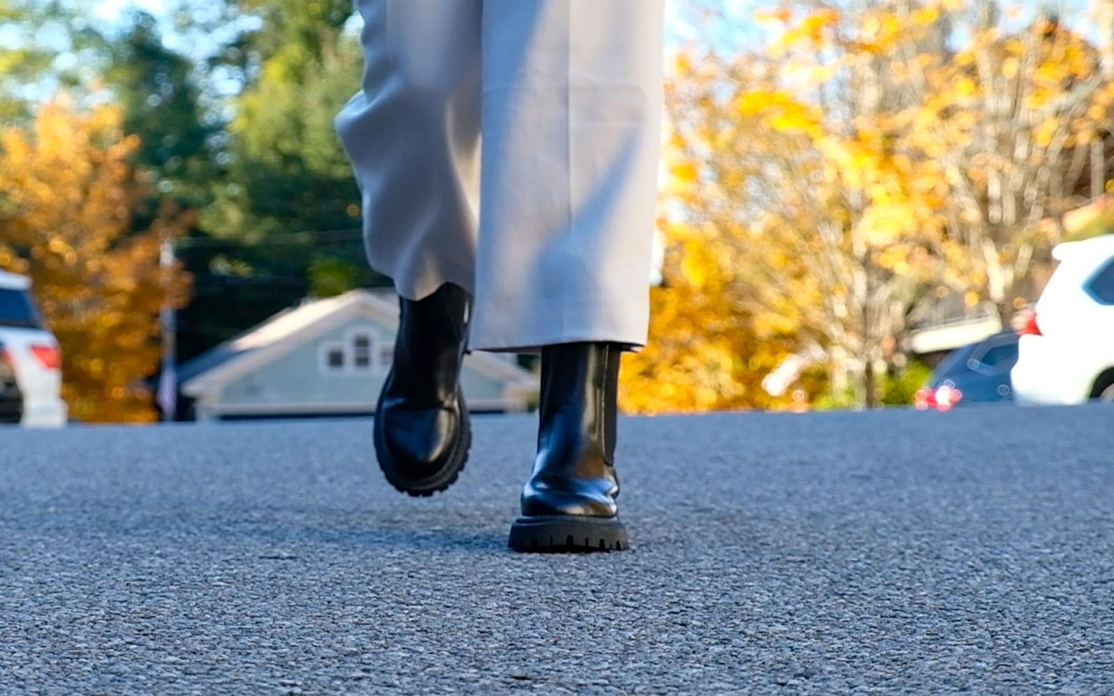 Black chunky boots and cream trousers walking across rough asphalt, autumn trees blurred behind