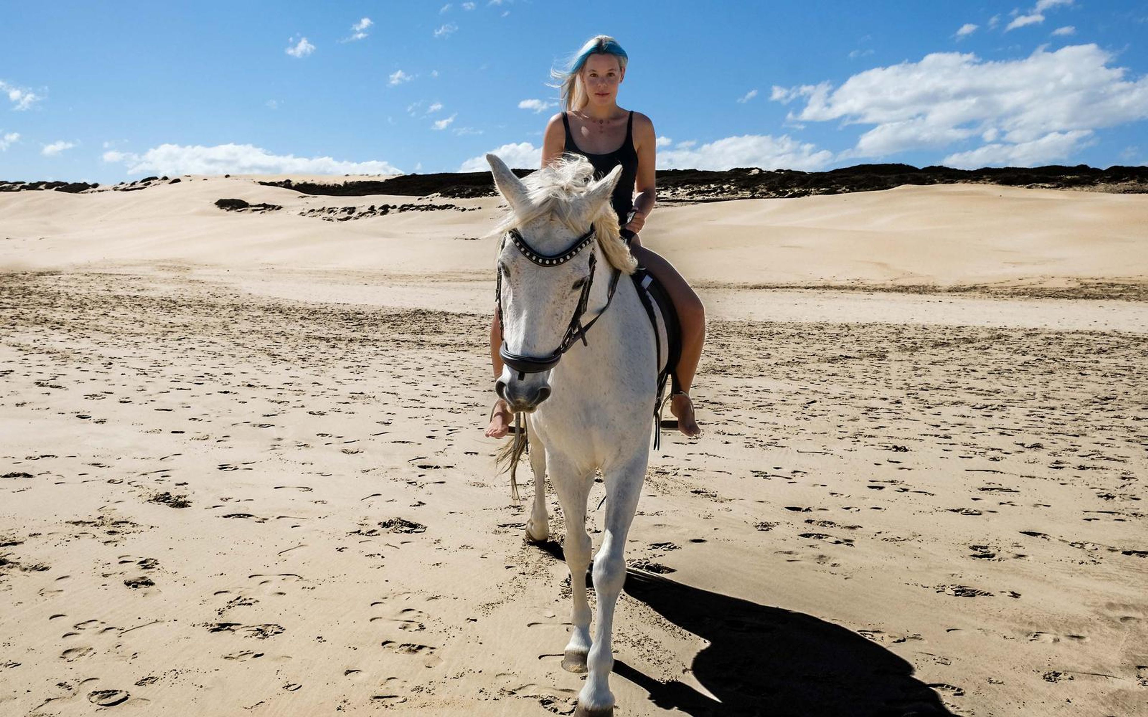 White horse and rider walking toward camera on wet sand, blue sky with scattered clouds
