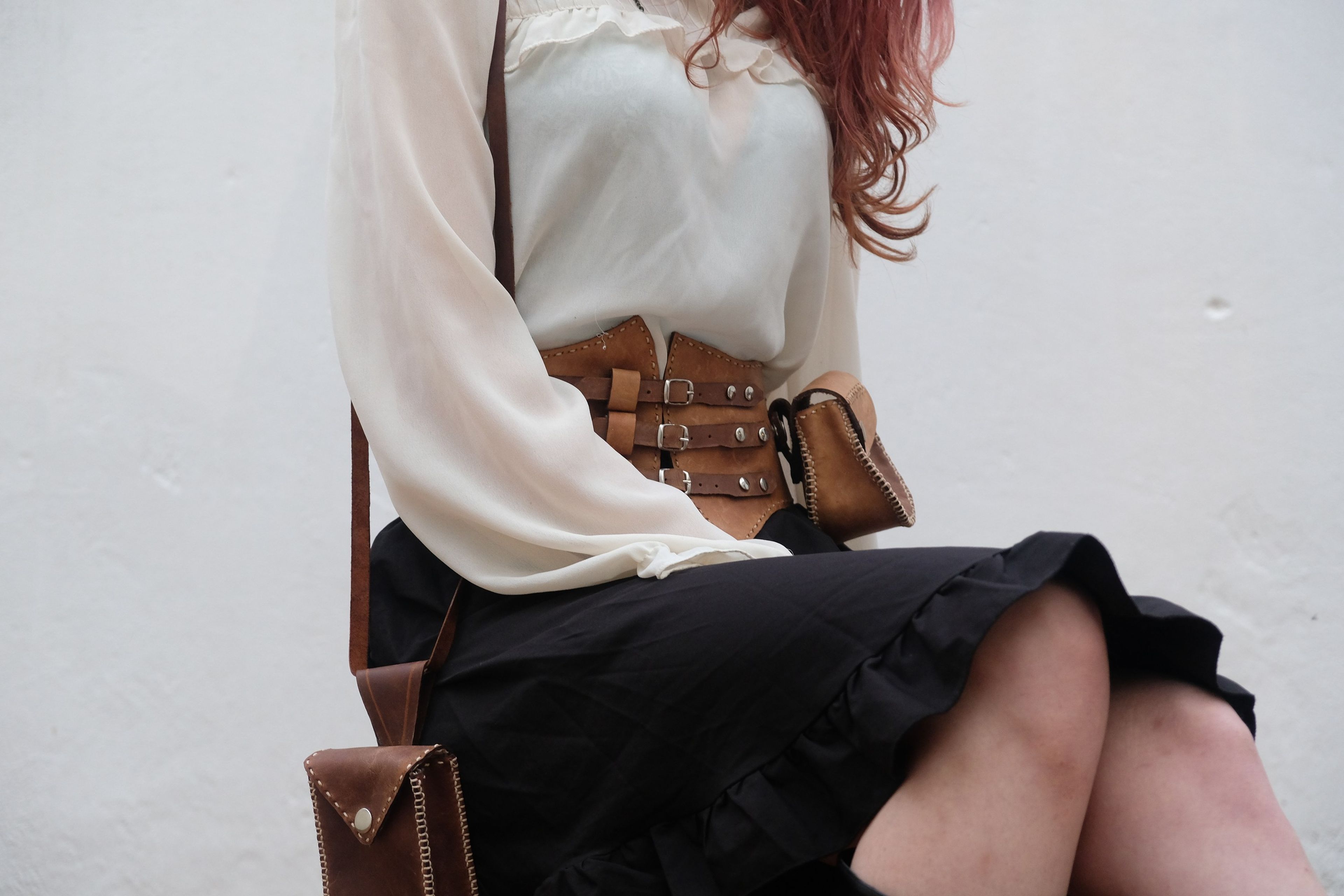 Woman seated in a cream blouse and black skirt wearing a leather corset belt and pouch with a shoulder bag