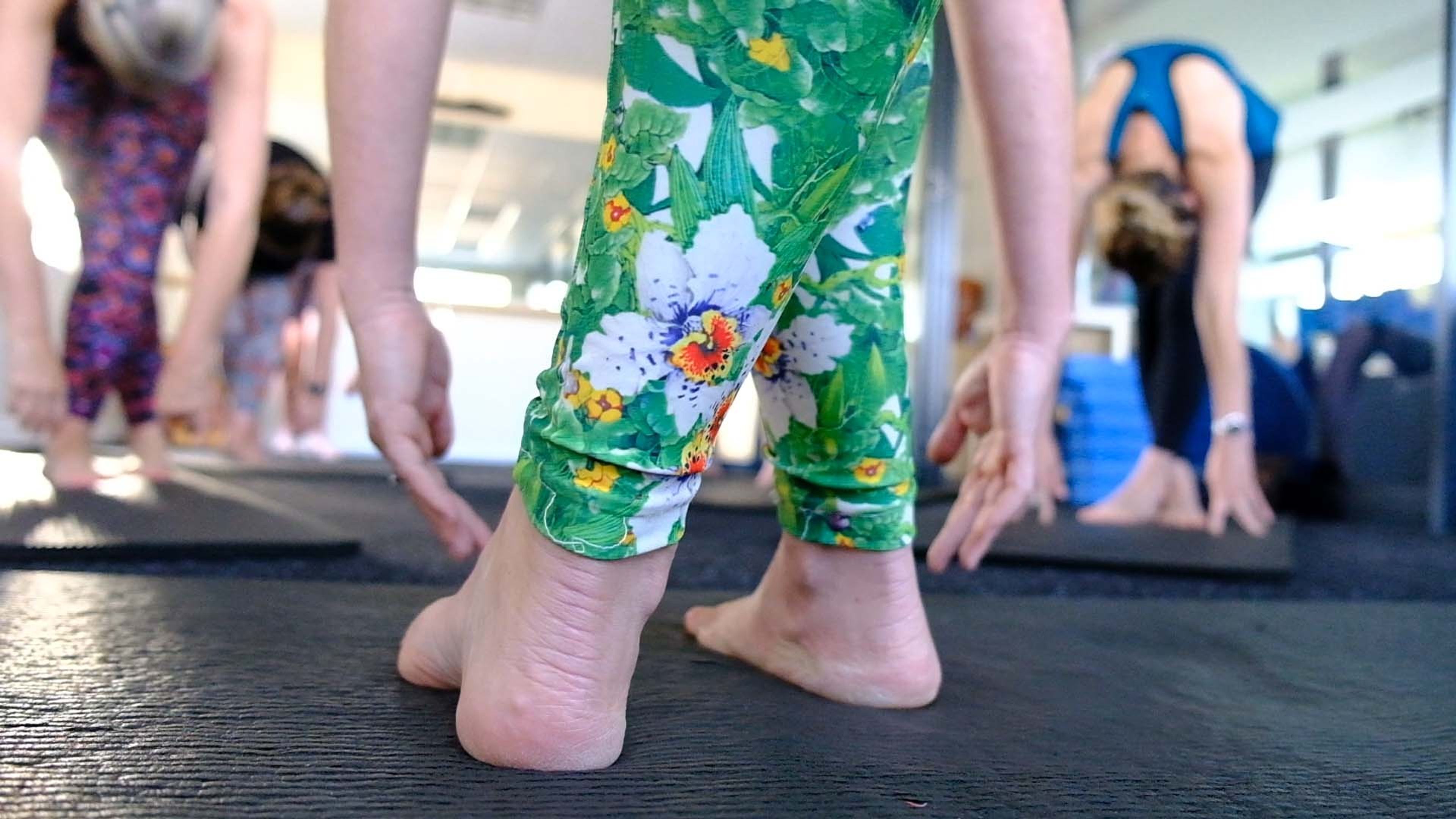 Close-up of bare feet and floral green leggings as a client folds forward on a mat, other class members bending behind